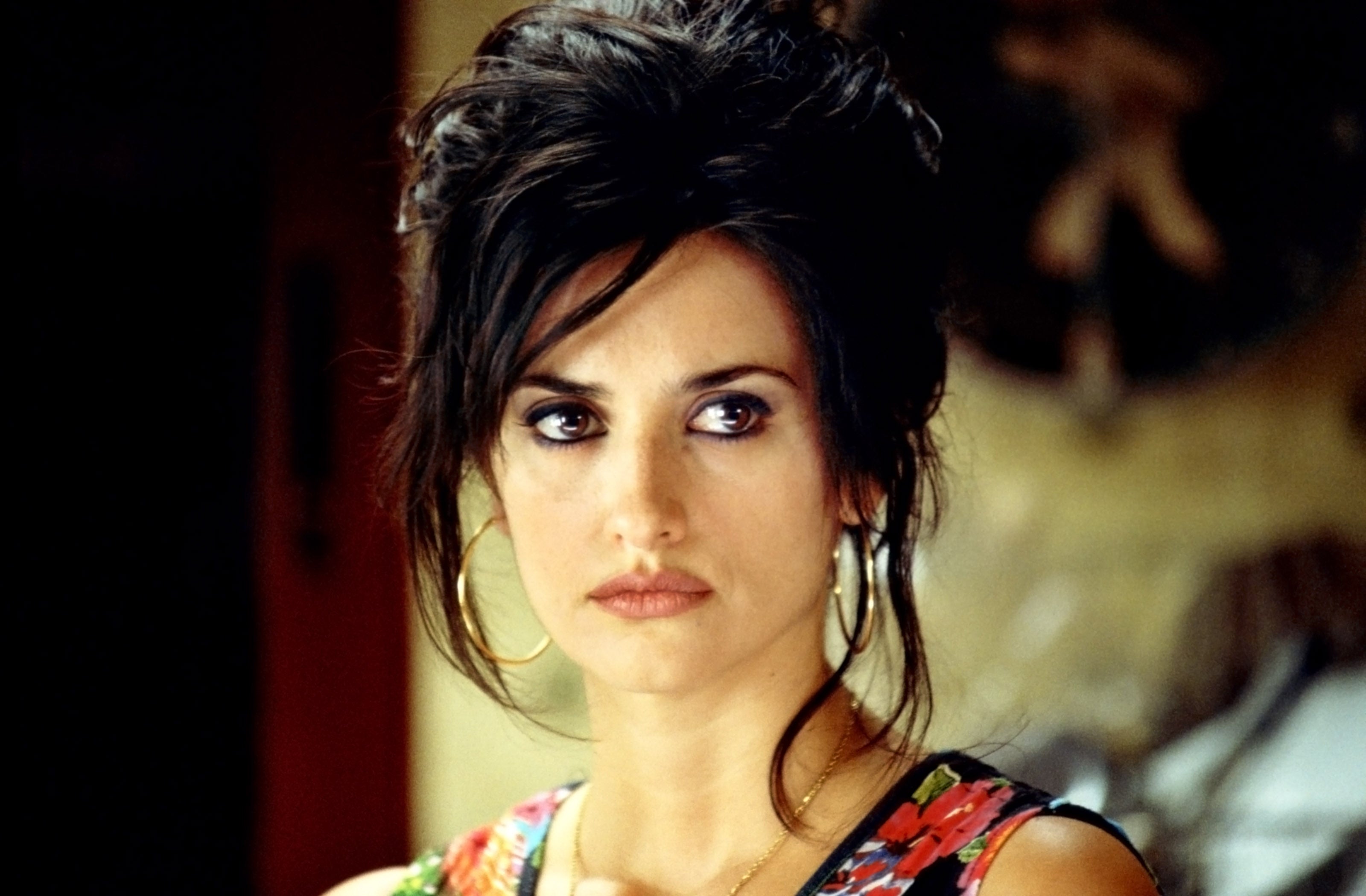 Penelope Cruz with intricate updo hairstyle, wearing a floral-patterned top and hoop earrings, gazing thoughtfully