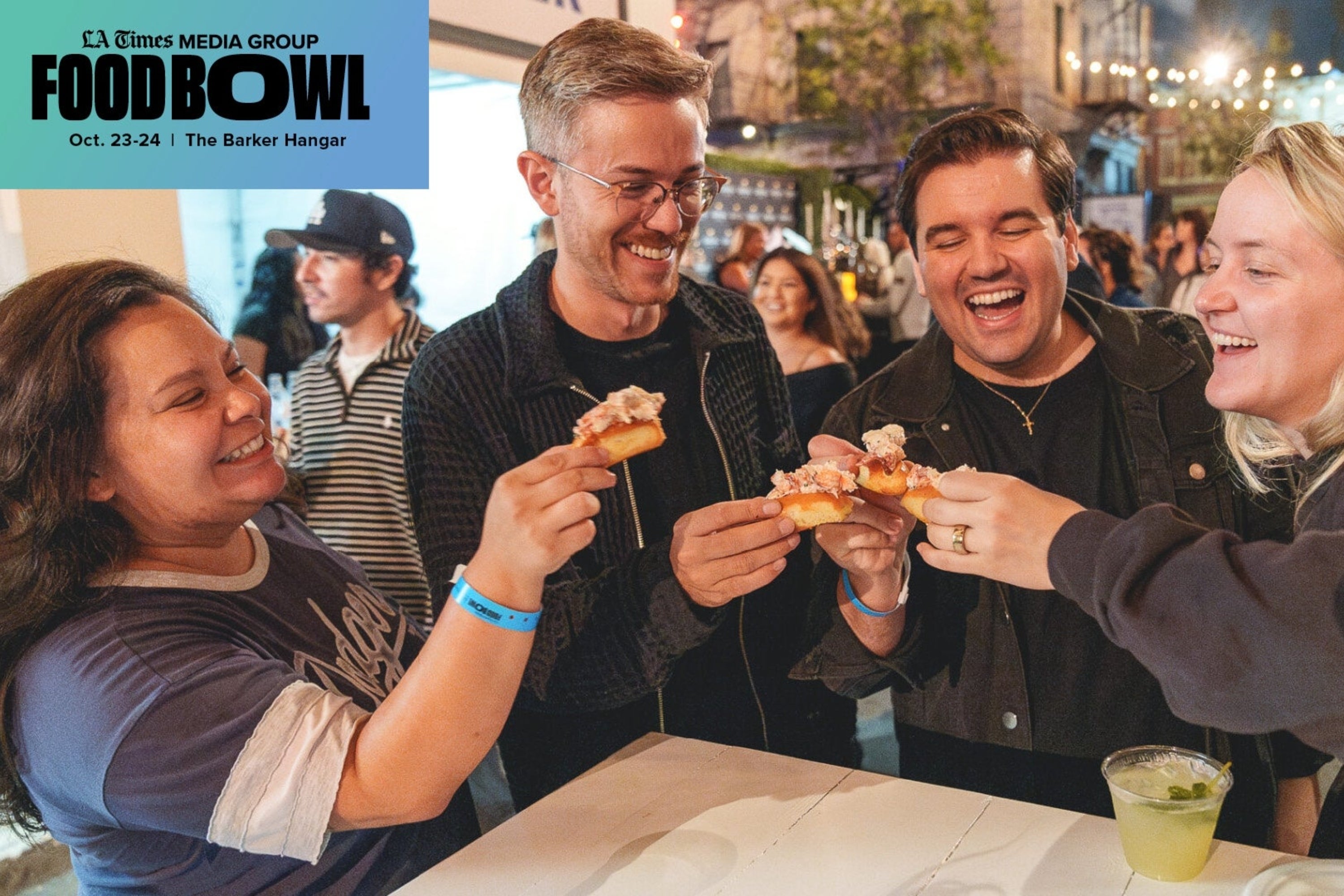 People enjoying appetizers at a food event, holding small bites and smiling together, with a drink on the table. FoodBowl event banner visible