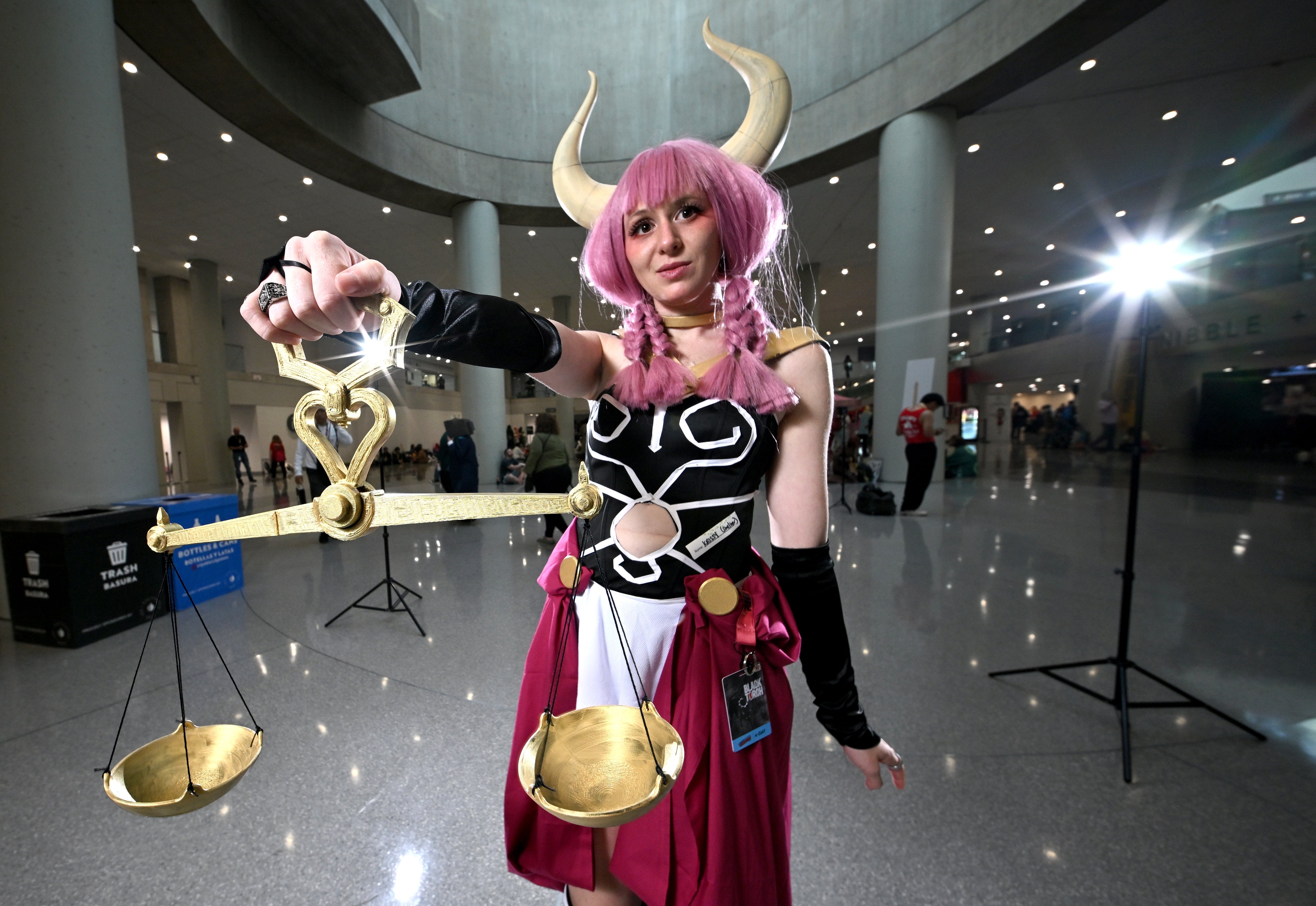 Person in a pink wig and elaborate cosplay costume holding a scale prop, posing in a convention center