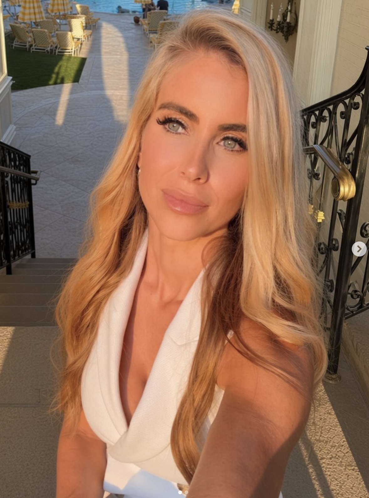 Person taking a selfie outdoors, wearing an elegant sleeveless outfit, with styled agelong hair. Background includes a terrace and distant water