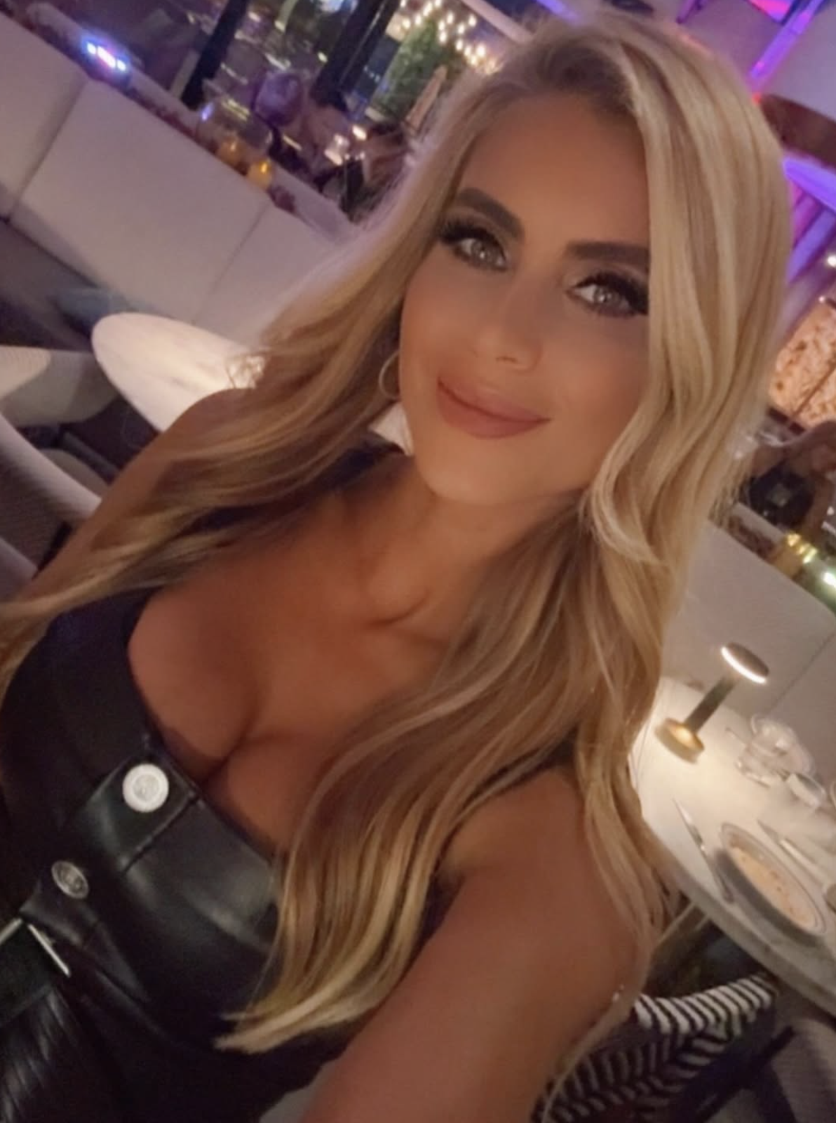 Person smiling successful a restaurant, wearing a sleeveless leather apical with fastener details. Tables and brushed lighting disposable successful the background