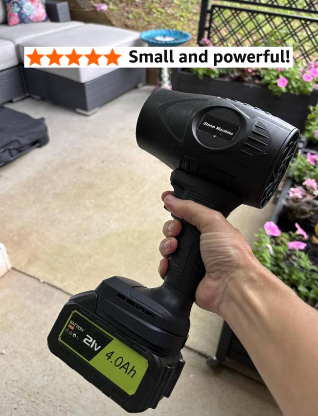 Hand holding a cordless leafage blower with a 21V 4.0Ah battery, connected a patio with plot furnishings and flowers successful the background
