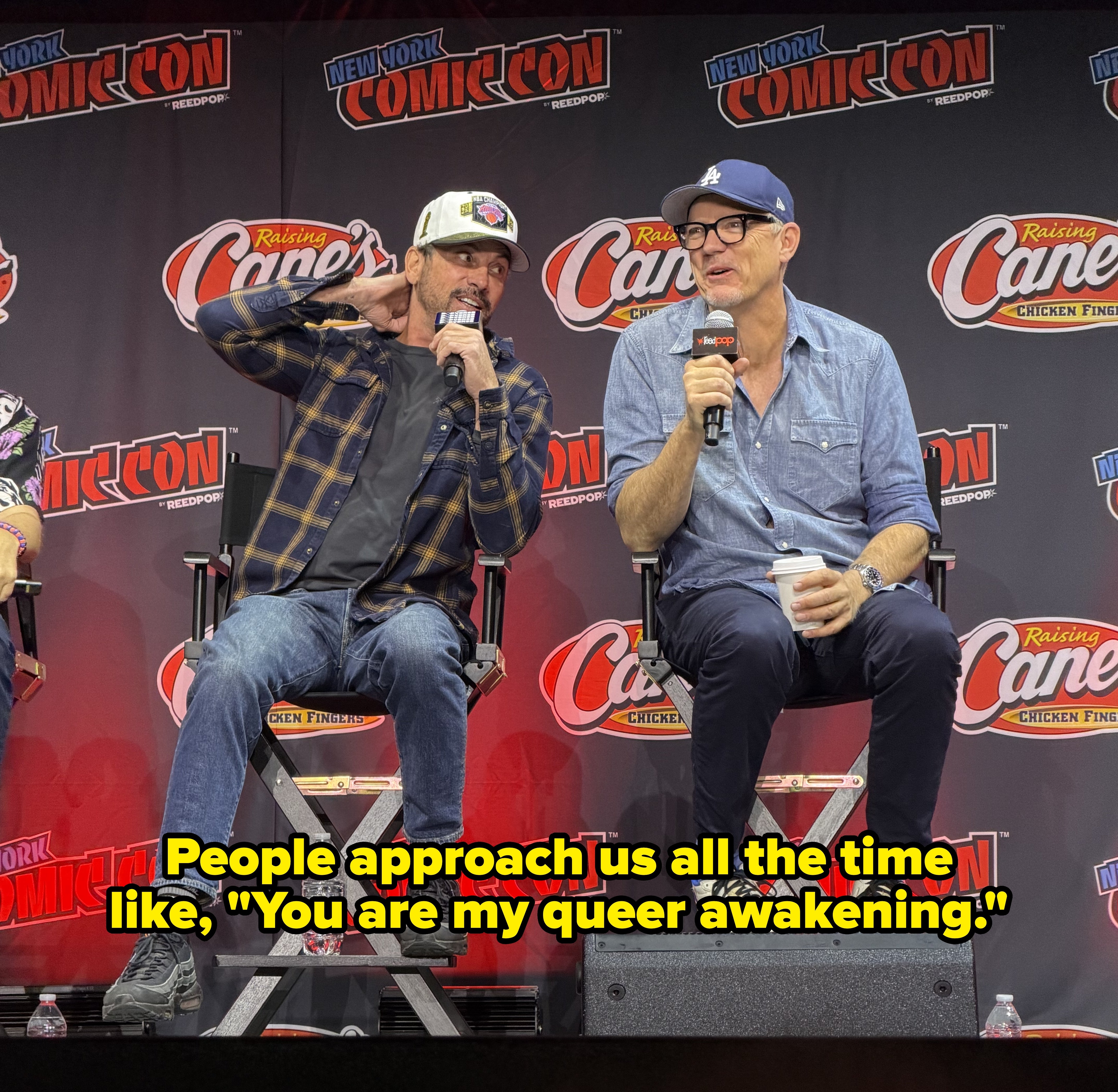 Two people sitting and speaking on stage at New York Comic Con, one in casual wear with a cap, the other in a shirt and glasses holding a cup