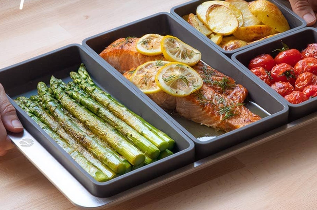 Divided baking tray with cooked salmon, asparagus, roasted potatoes, and cherry tomatoes, illustrating organized meal prep