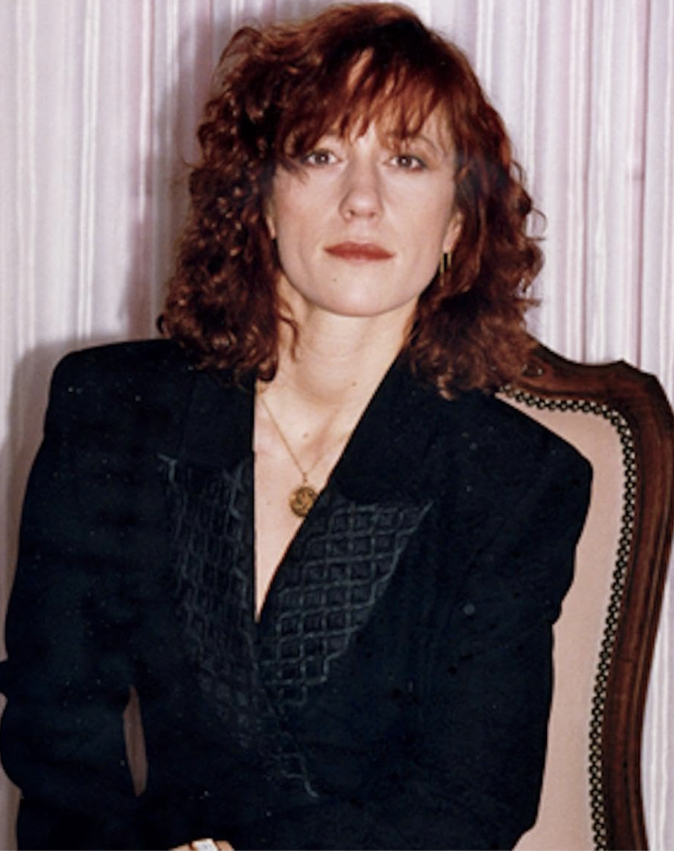 Person with wavy hair sits in a chair, wearing a dark textured blazer with a pendant necklace