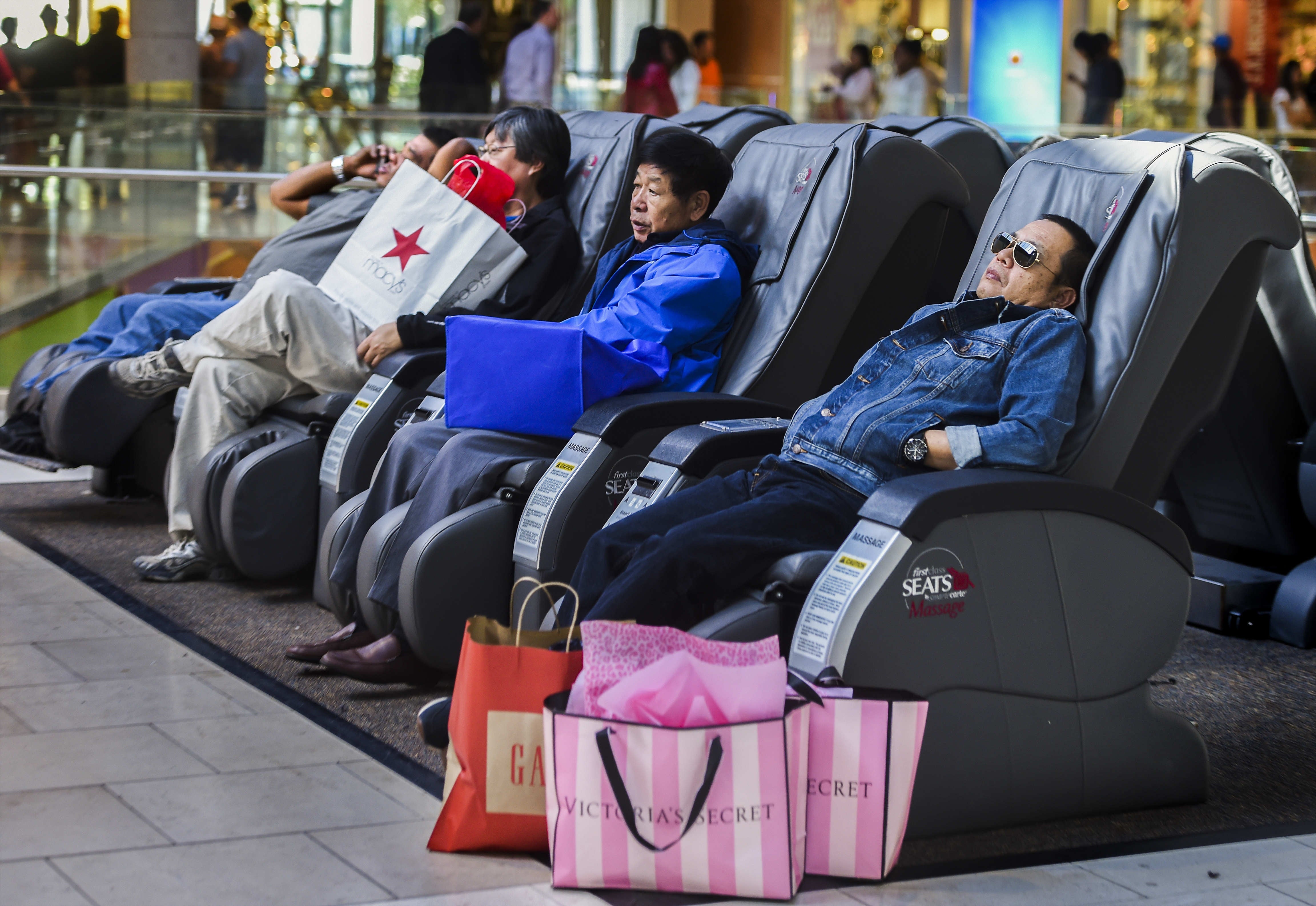 People relaxing in massage chairs at a mall, surrounded by shopping bags
