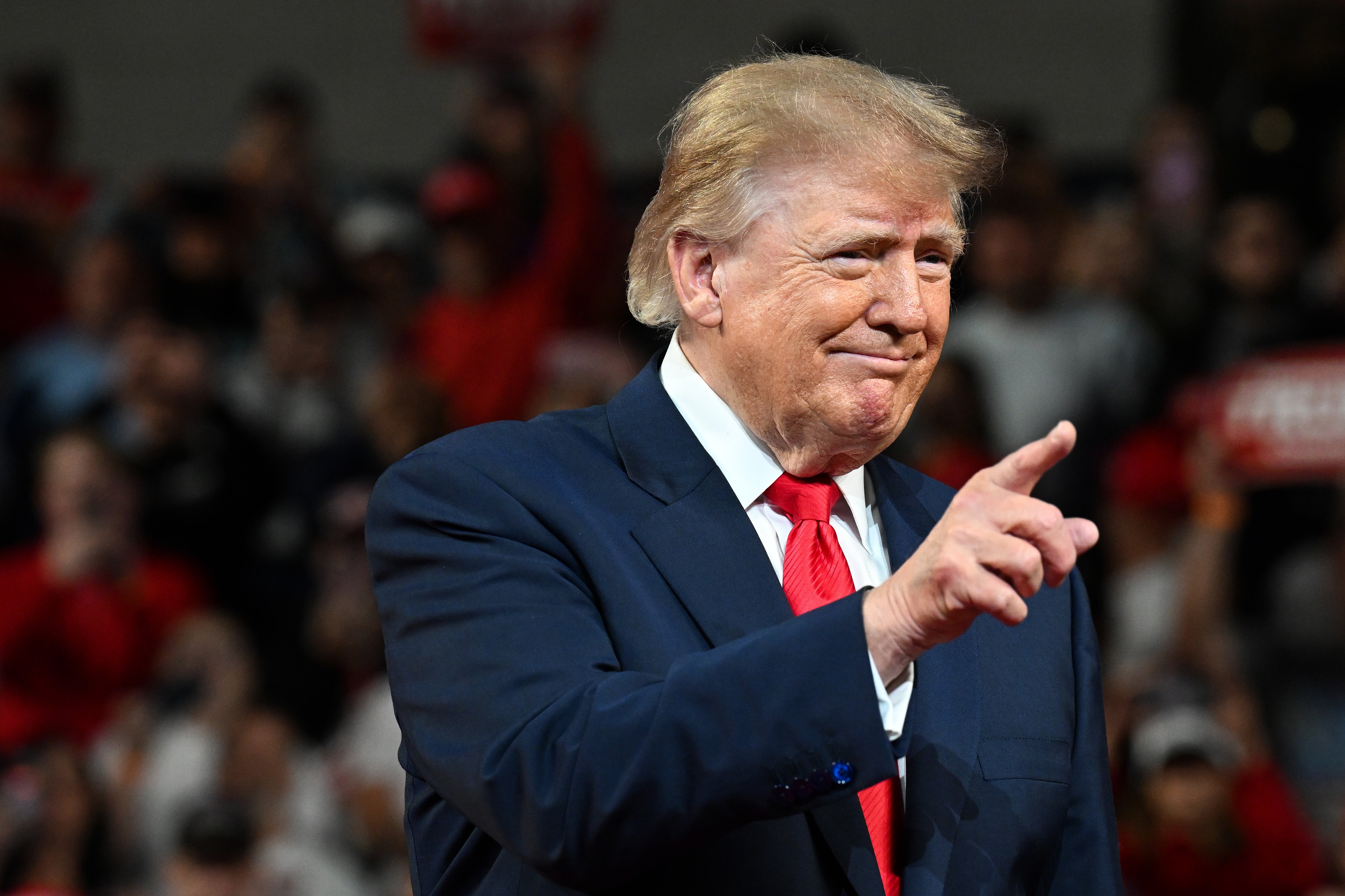 A person in a suit gestures with a finger, standing in front of a crowd at an event