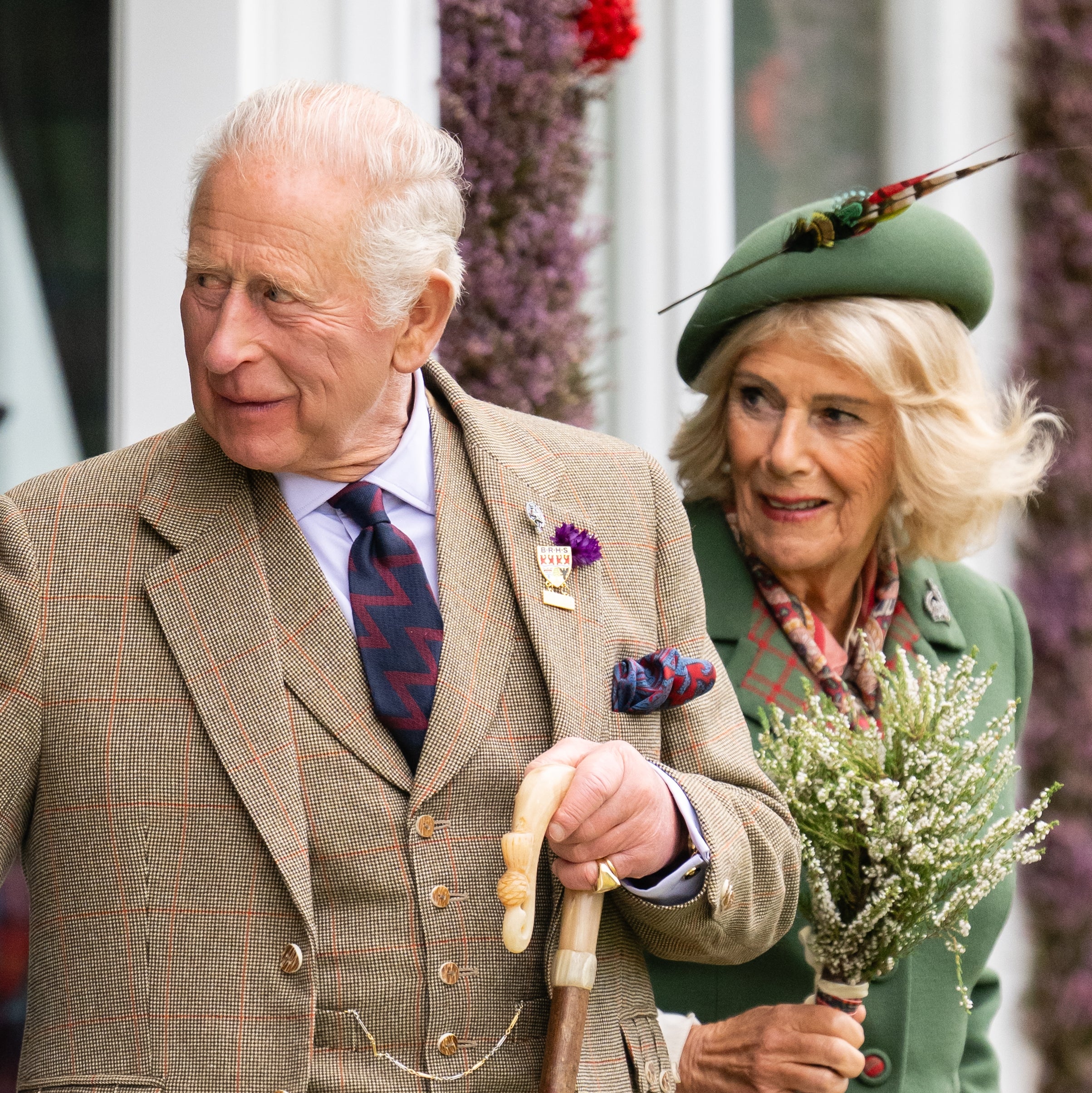 Two older adults in plaid attire, one with a cane, and the other holding a small bouquet, smiling and looking to the side