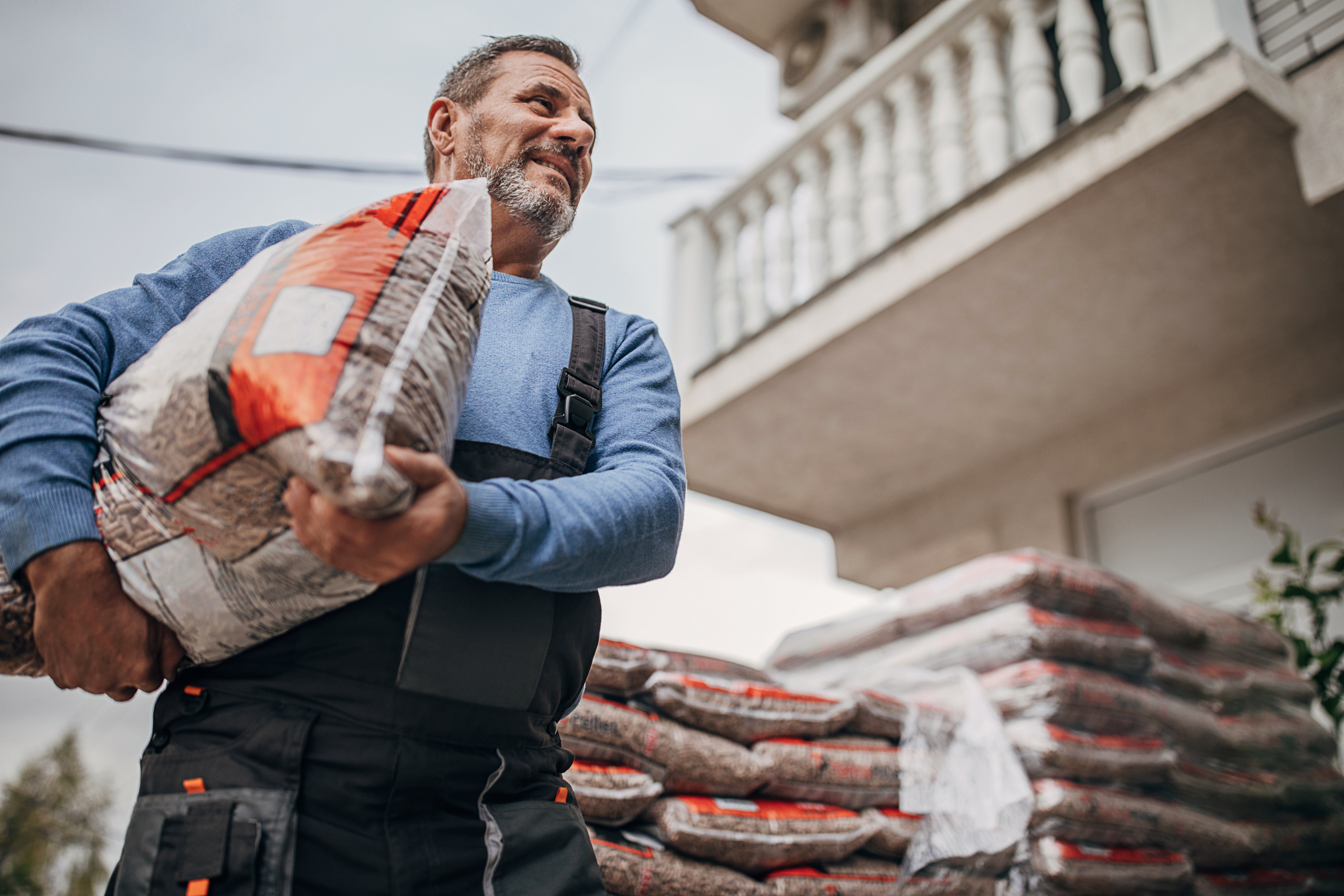 Man in work clothes carrying a bag of construction material at a job site, with more bags stacked in the background