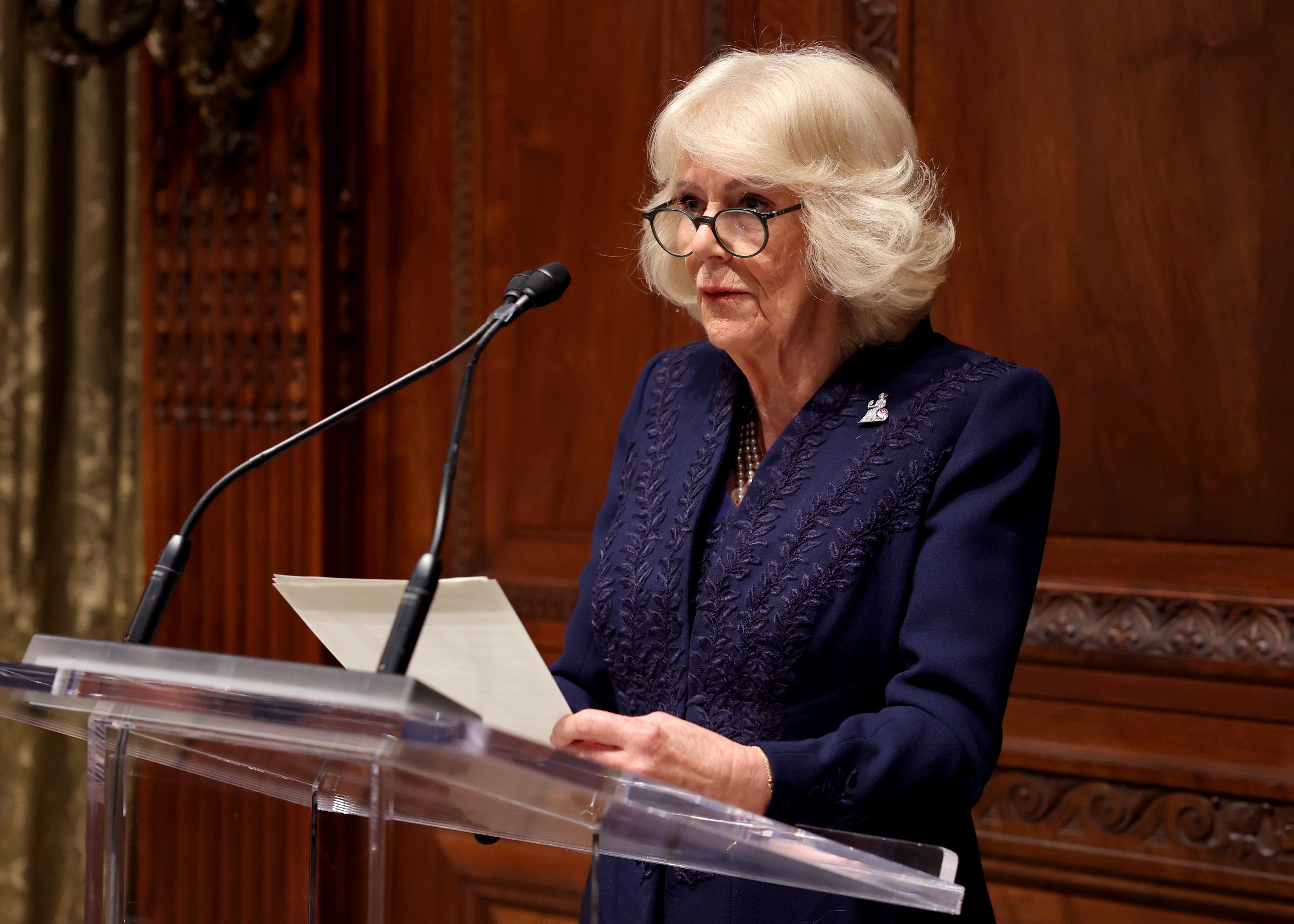 An individual in formal attire speaks at a podium, holding a paper
