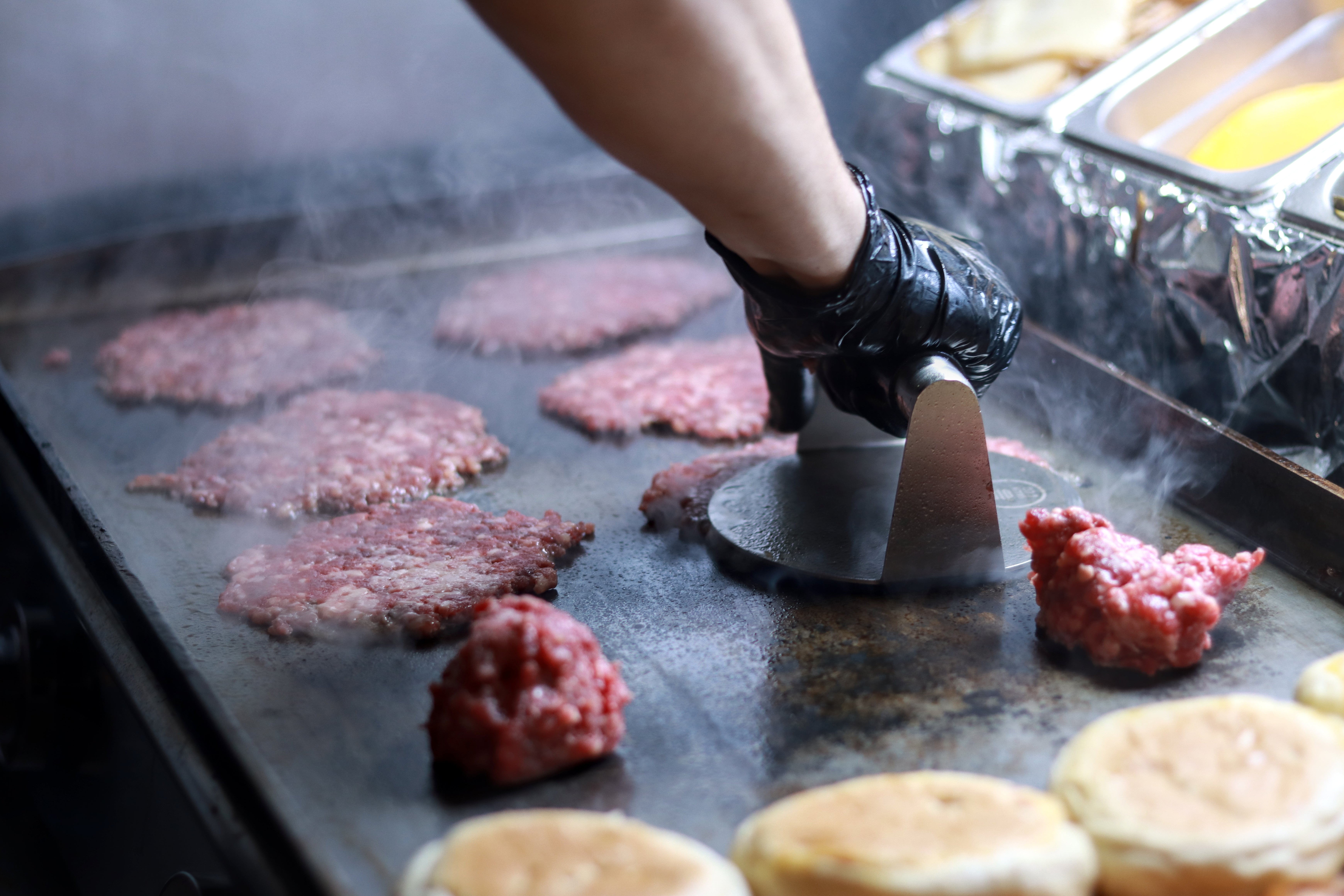 Person wearing a glove presses ground beef patties onto a grill, with buns in the foreground, in a kitchen setting