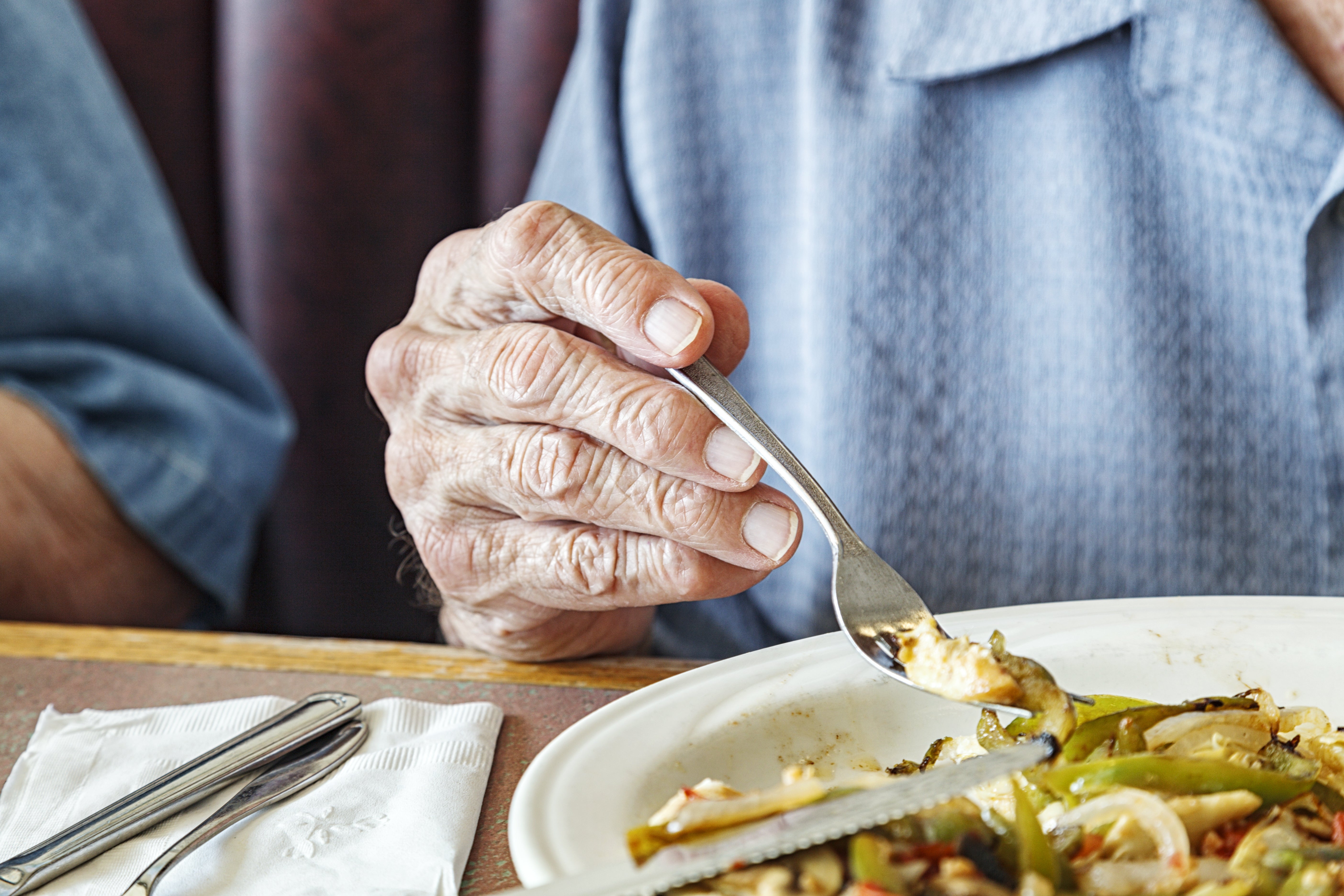 Elderly person using a fork to eat from a plate of vegetables at a table, displaying a focused grip on the utensil