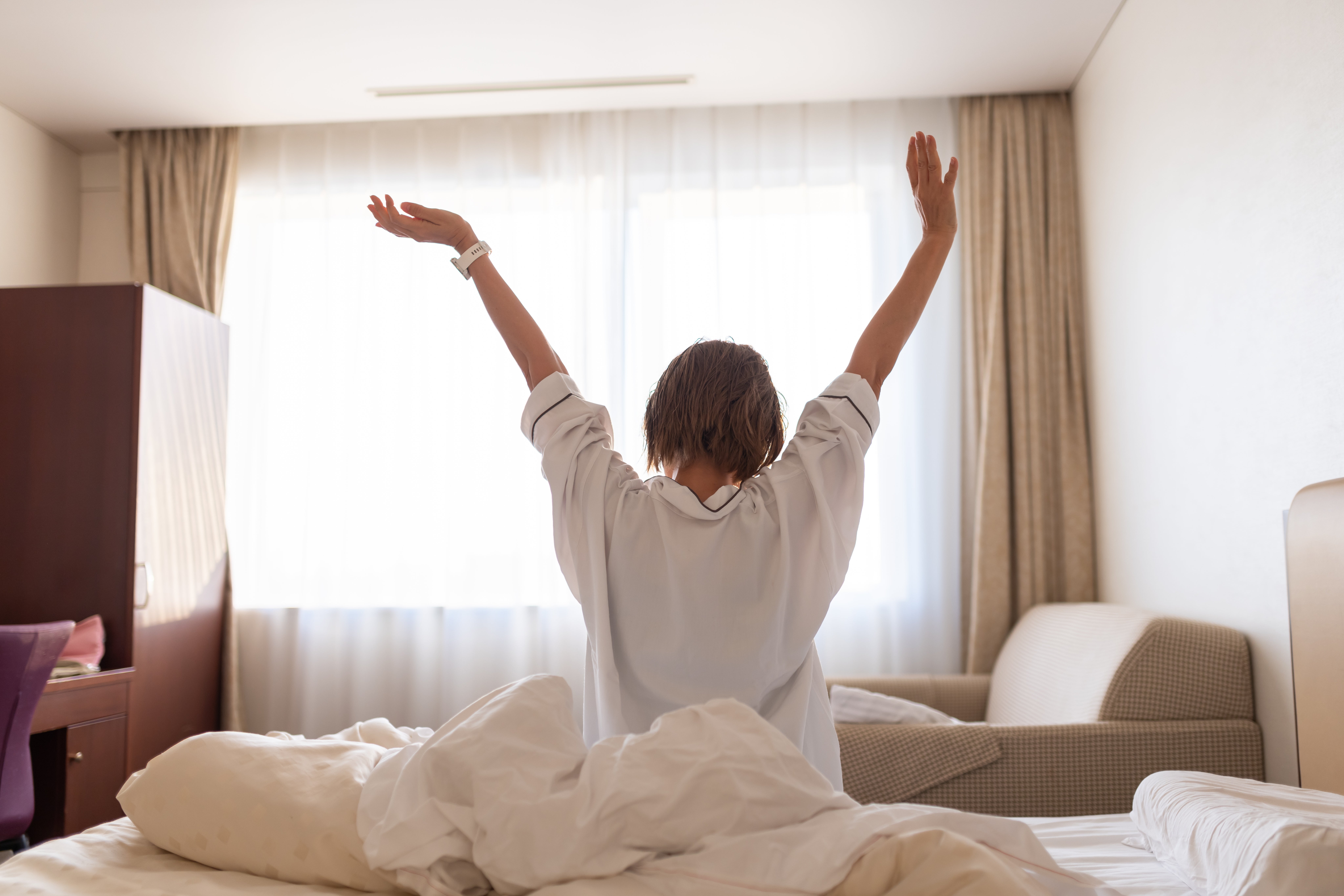 Person stretching in bed, facing a window with sheer curtains, suggesting a morning scene in a cozy room