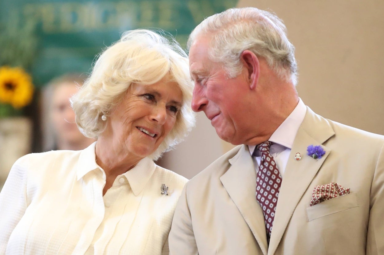 Two individuals smiling warmly at each other; one wears a light suit with a floral pin, and the other wears a classic collared shirt