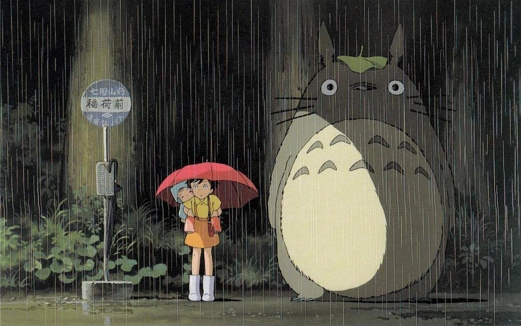 Two characters stand in the rain at a bus stop; a giant creature with a leaf on its head and a child with an umbrella and younger companion