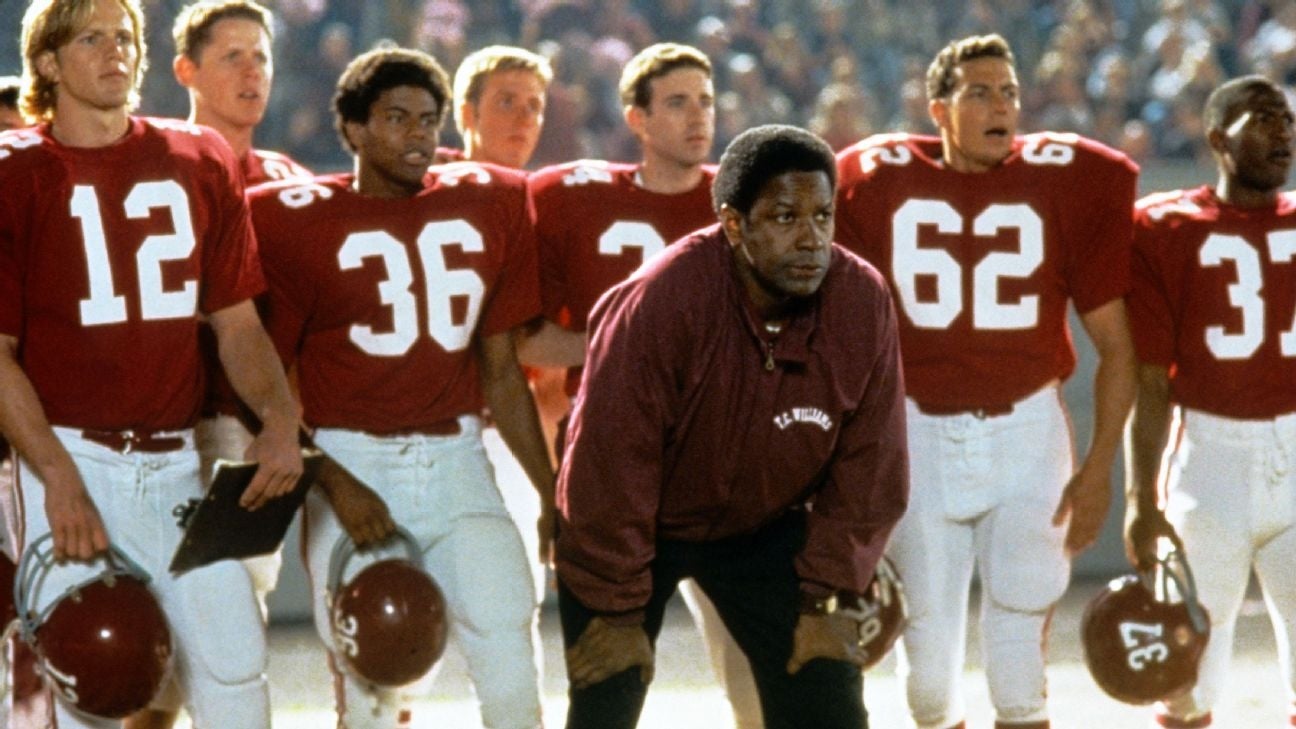 Football players and a coach stand on the sidelines during a game, focused and intent. Players wear numbered jerseys and helmets