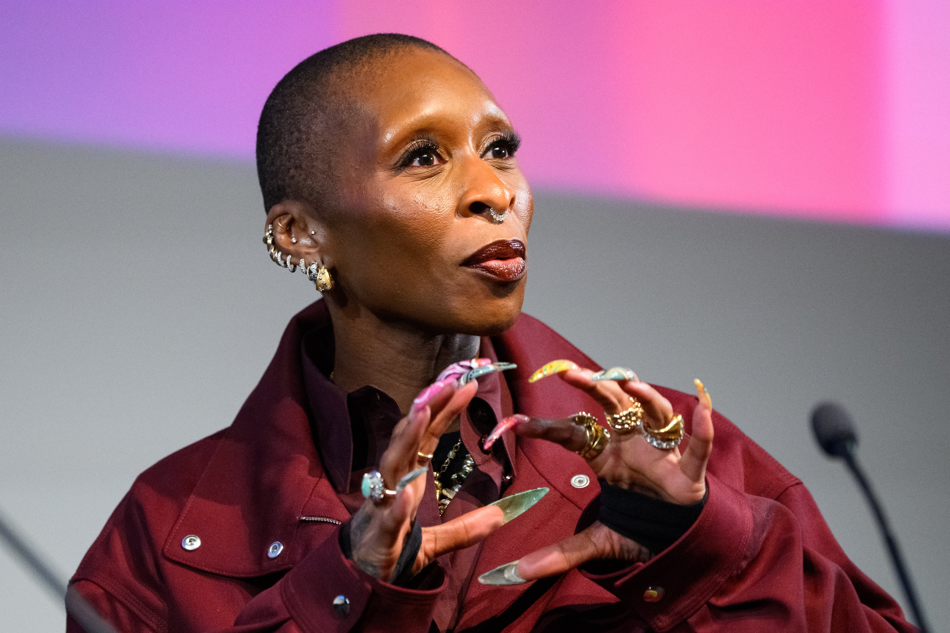Person with a stylish shaved head wearing a bold layered outfit, gesturing expressively while speaking at an event