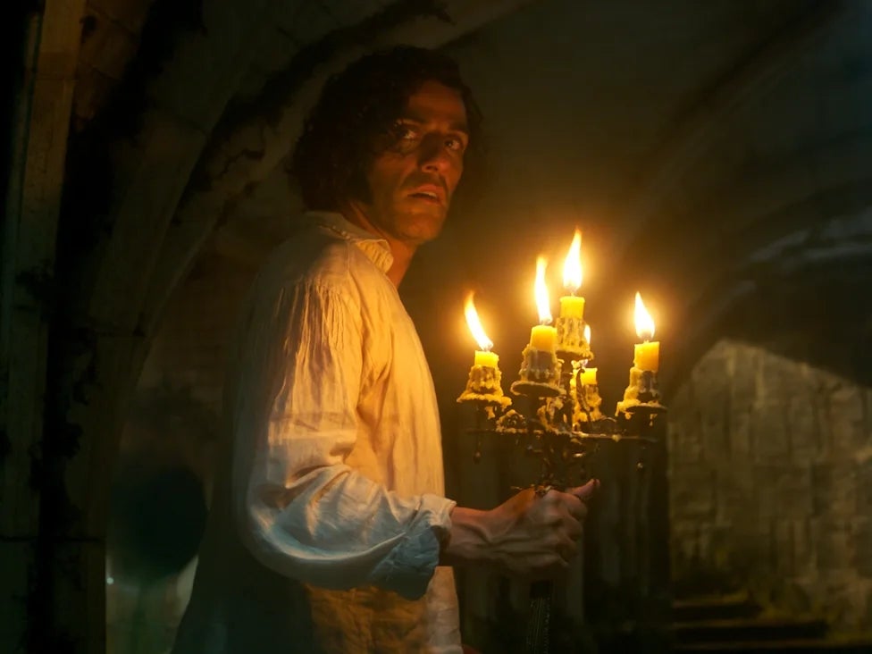 Person holding a candelabra in a dim, stone-walled setting, wearing a loose shirt, looking back. Scene suggests mystery or suspense