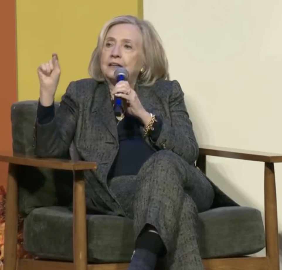 A person in a patterned suit is speaking into a microphone while seated, gesturing with one hand