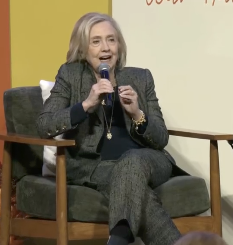 A person speaks into a microphone while sitting on a stage chair, wearing a matching blazer and pants, engaged in a public discussion