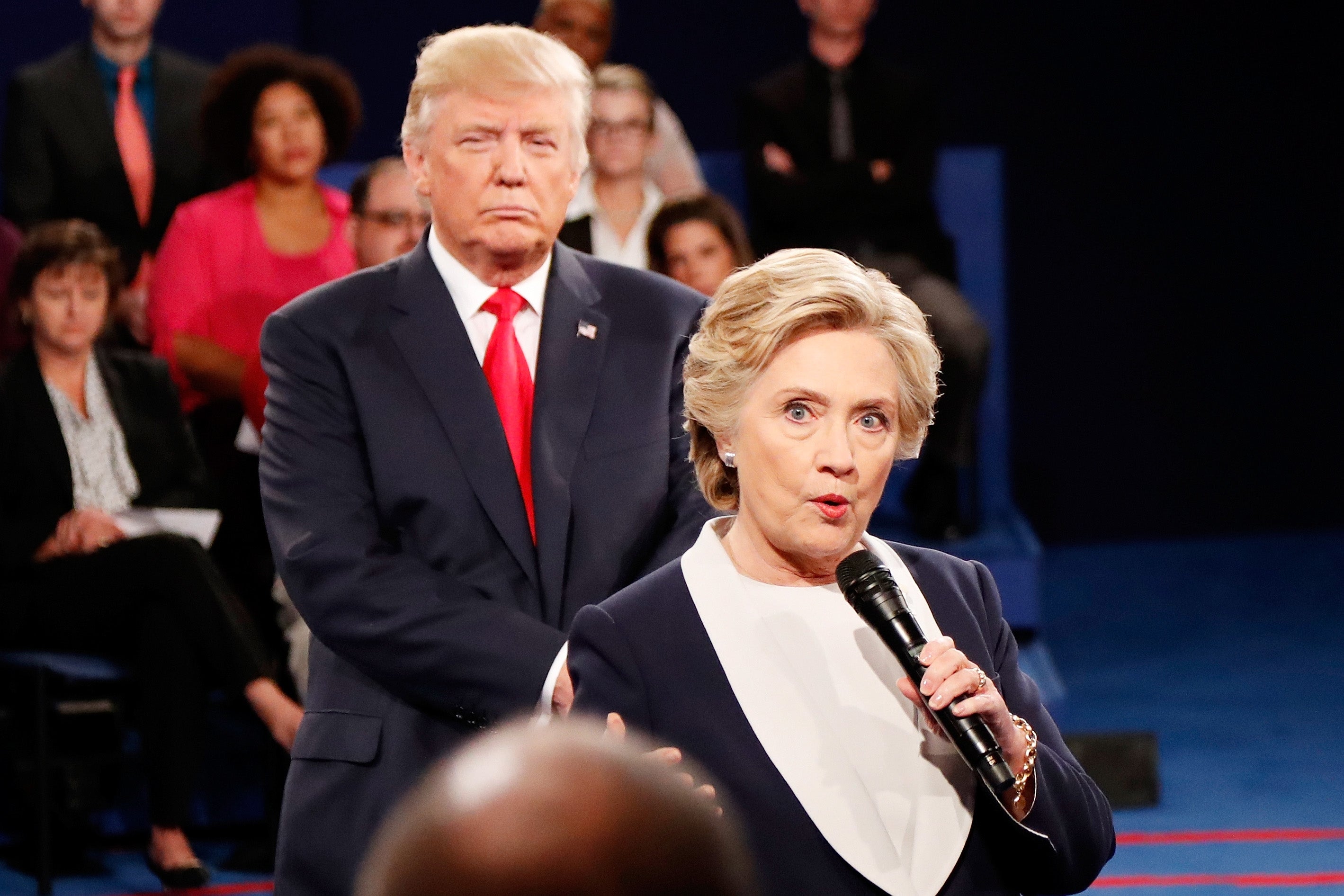 A woman and a man stand on a stage during a debate. The woman holds a microphone while addressing the audience