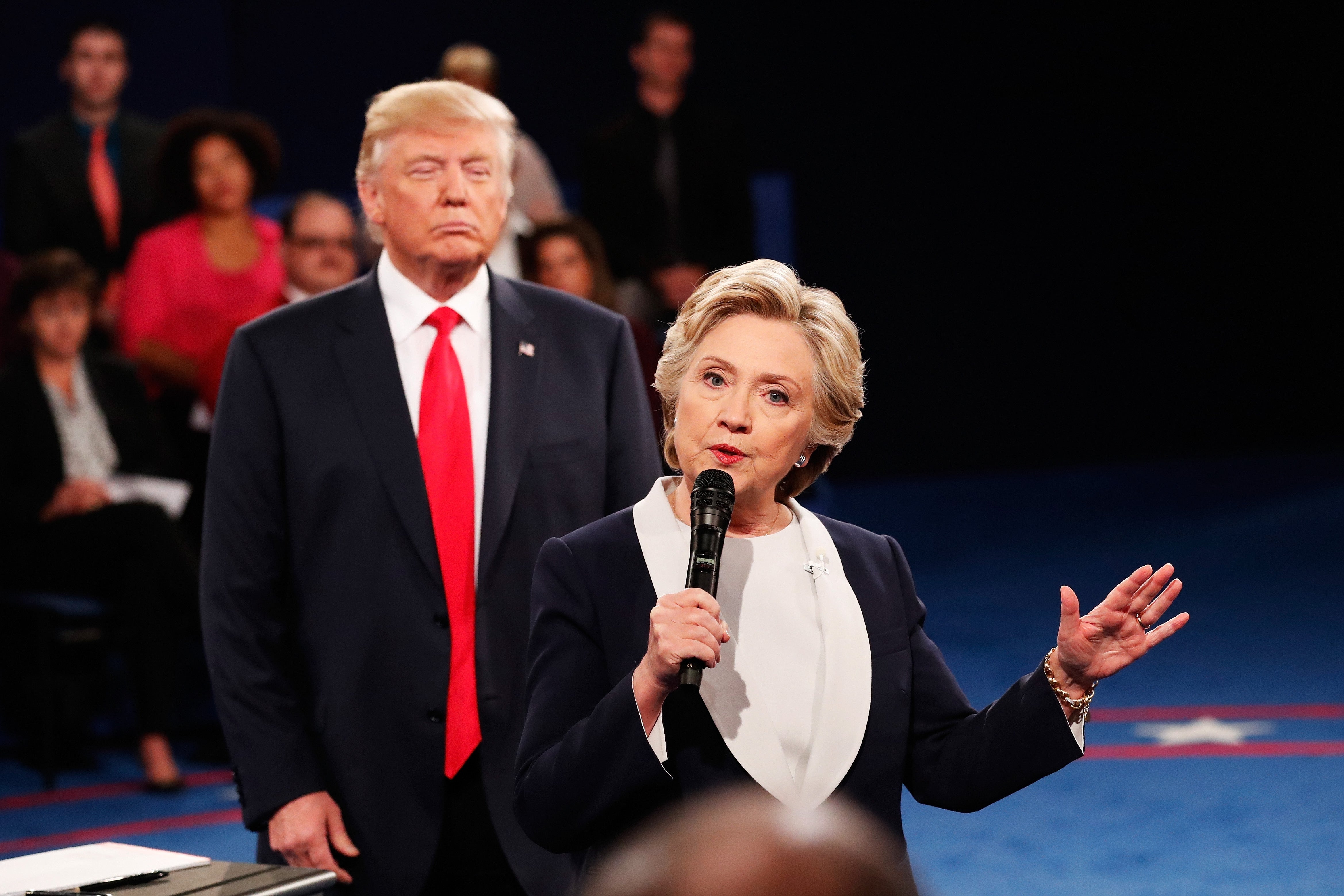 Two politicians standing on stage, one speaking into a microphone while the other stands behind, both in formal attire. Audience in the background