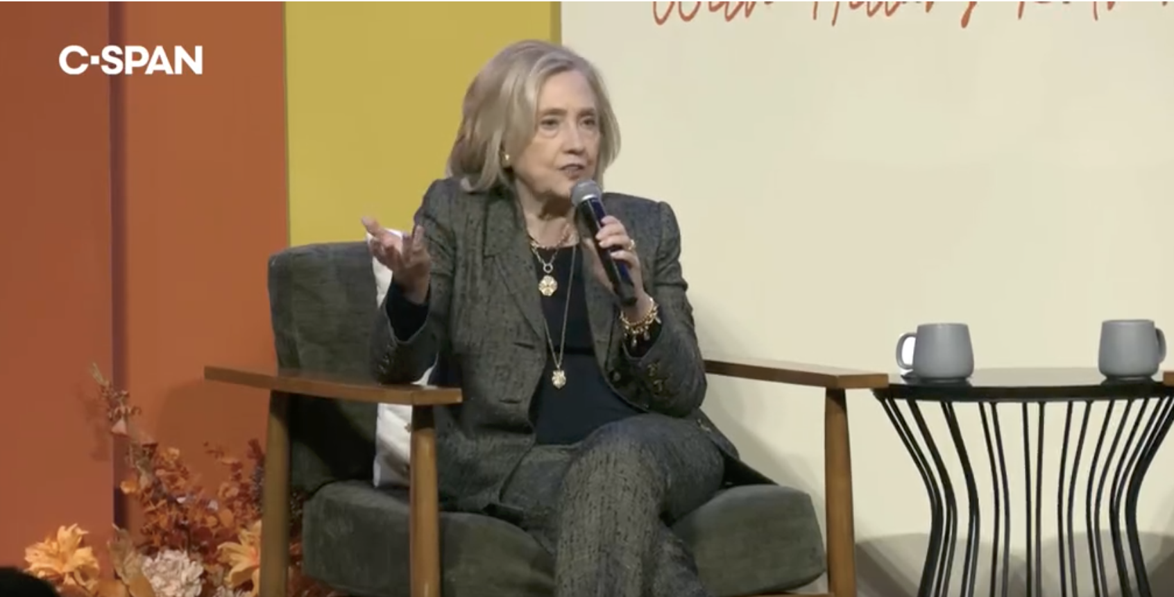 A person in a patterned suit speaks into a microphone while seated on a stage with two mugs on a table beside them