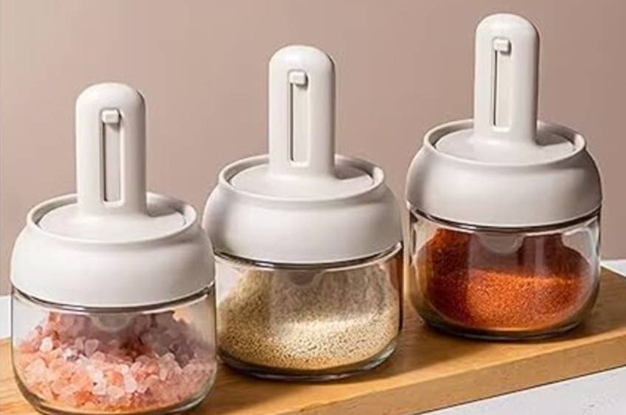 Three glass spice jars with white lids on a wooden tray, containing pink salt, grains, and red spice, suitable for kitchen organization