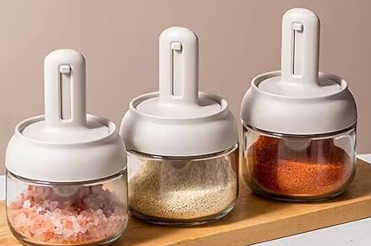 Three glass spice jars with white lids on a wooden tray, containing pink salt, grains, and red spice, suitable for kitchen organization