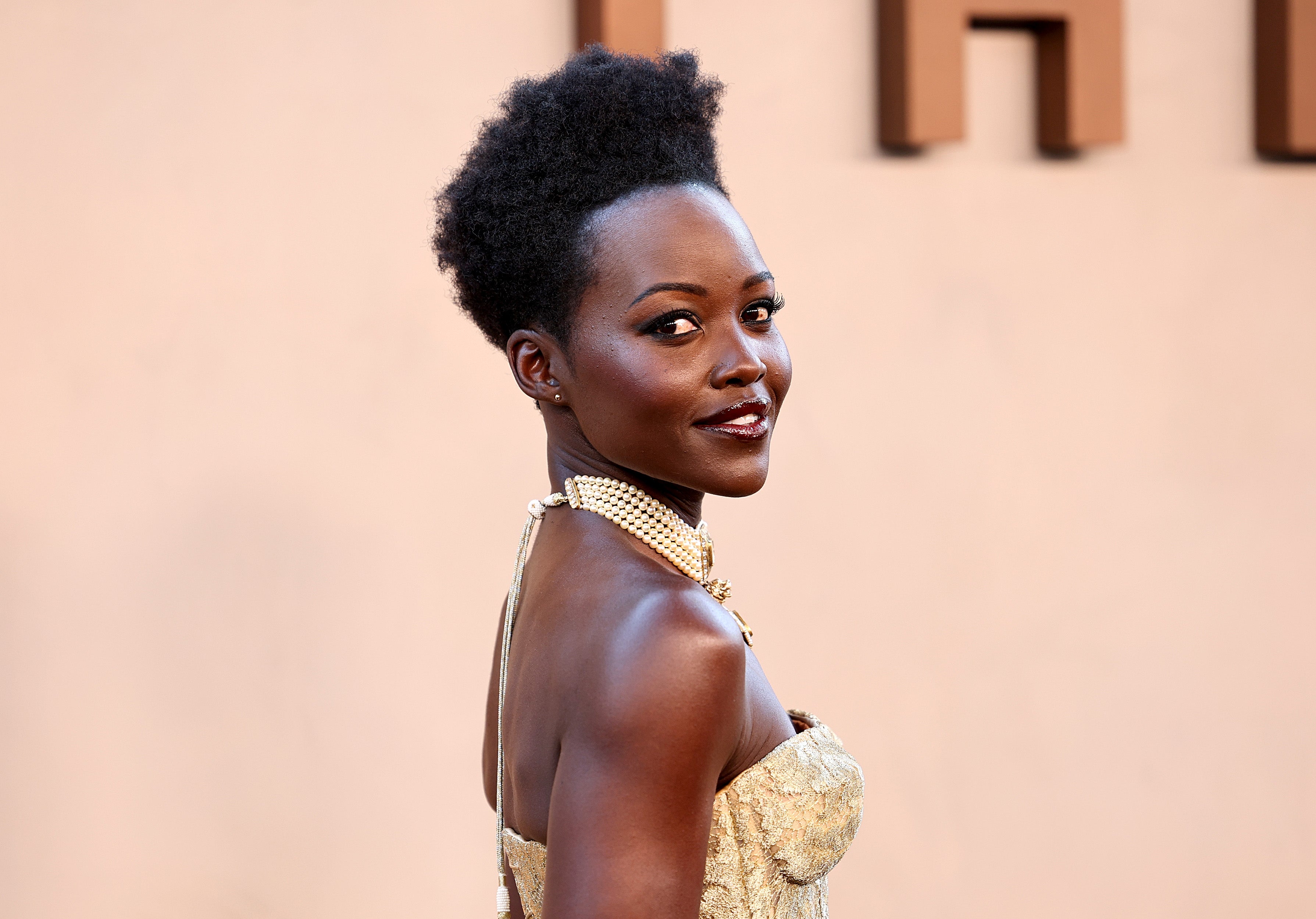 Person in elegant strapless dress, adorned with pearls, poses on a red carpet, exuding confidence