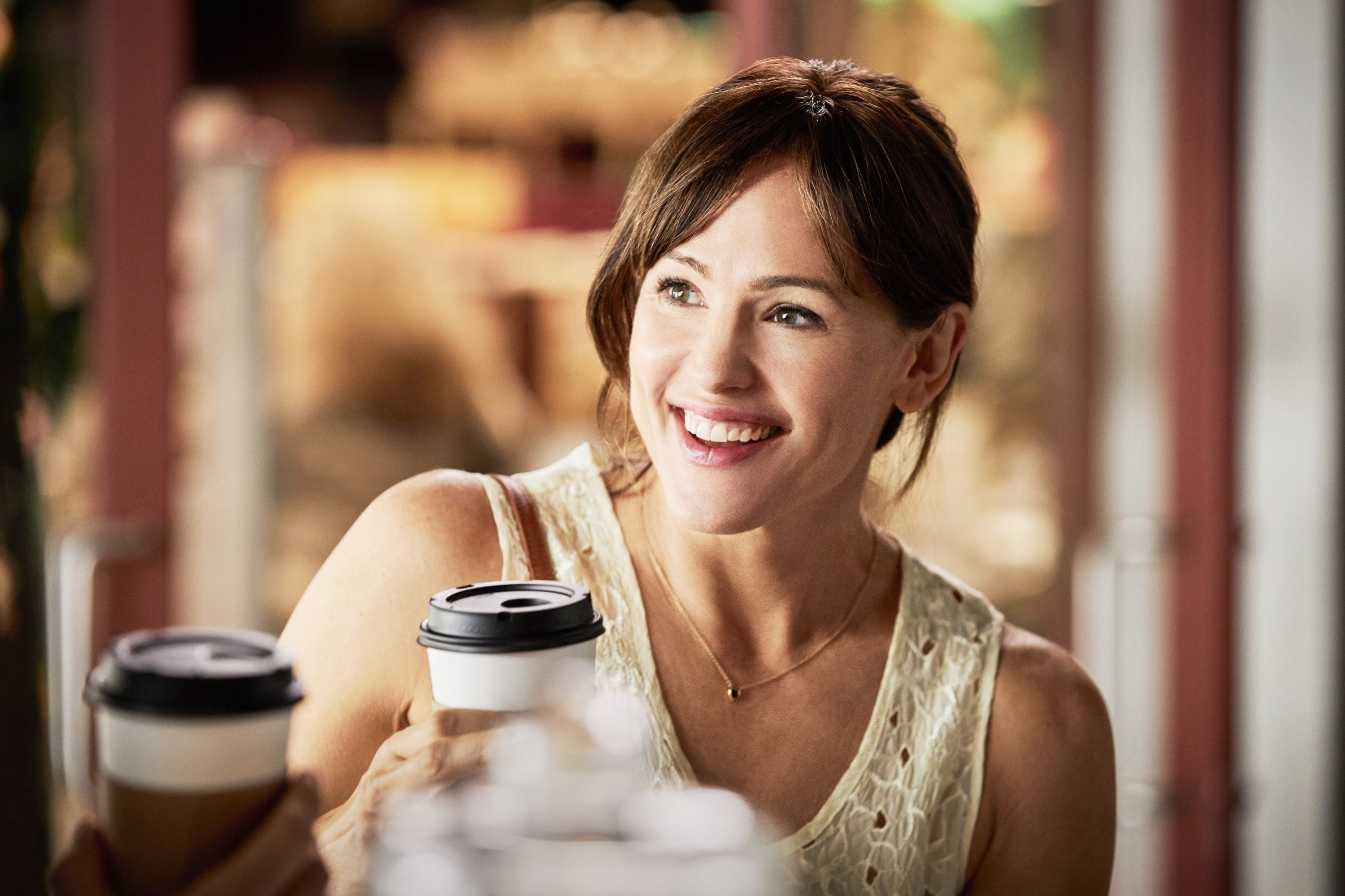 Person smiling while holding a takeaway coffee cup, wearing a sleeveless lace top, seated indoors