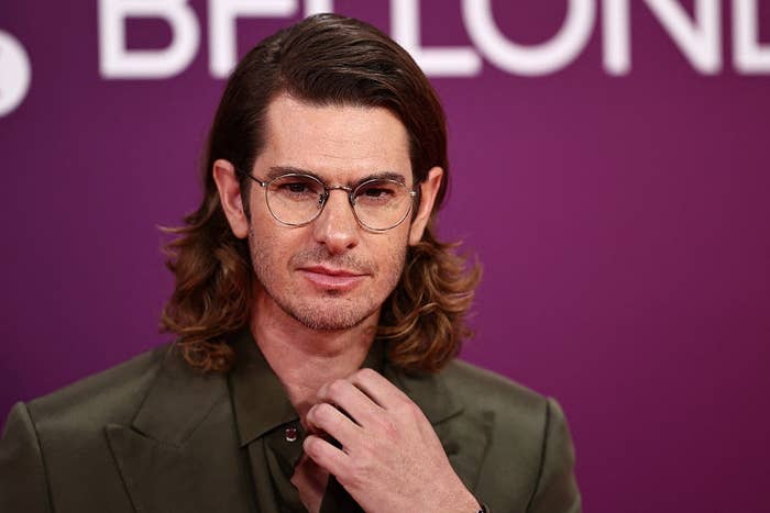 Person with long hair and glasses at an event, wearing a dark formal suit, posing with a thoughtful expression