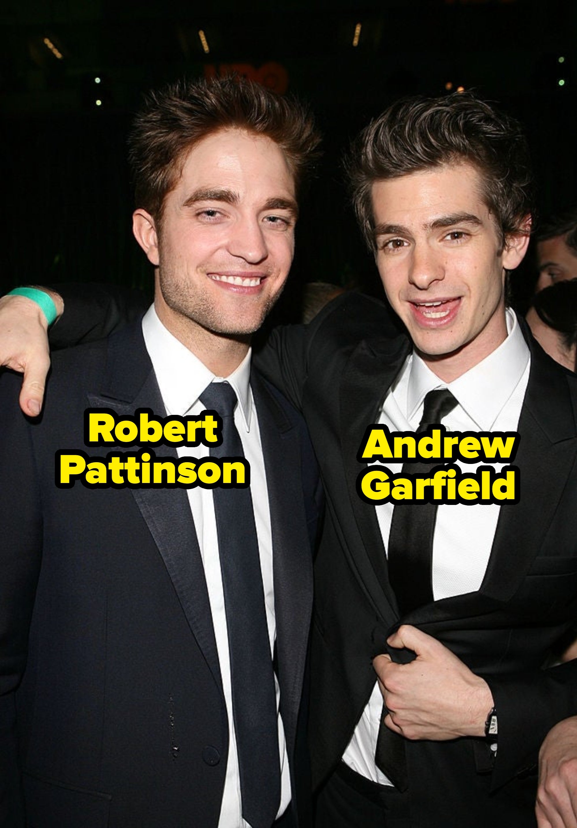 Two men in suits pose together at an event, one with his arm around the other. Both are smiling and dressed in classic formal attire