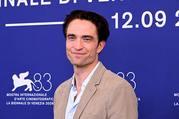 Actor in a beige suit and light shirt poses in front of a Venice Film Festival backdrop with a smile
