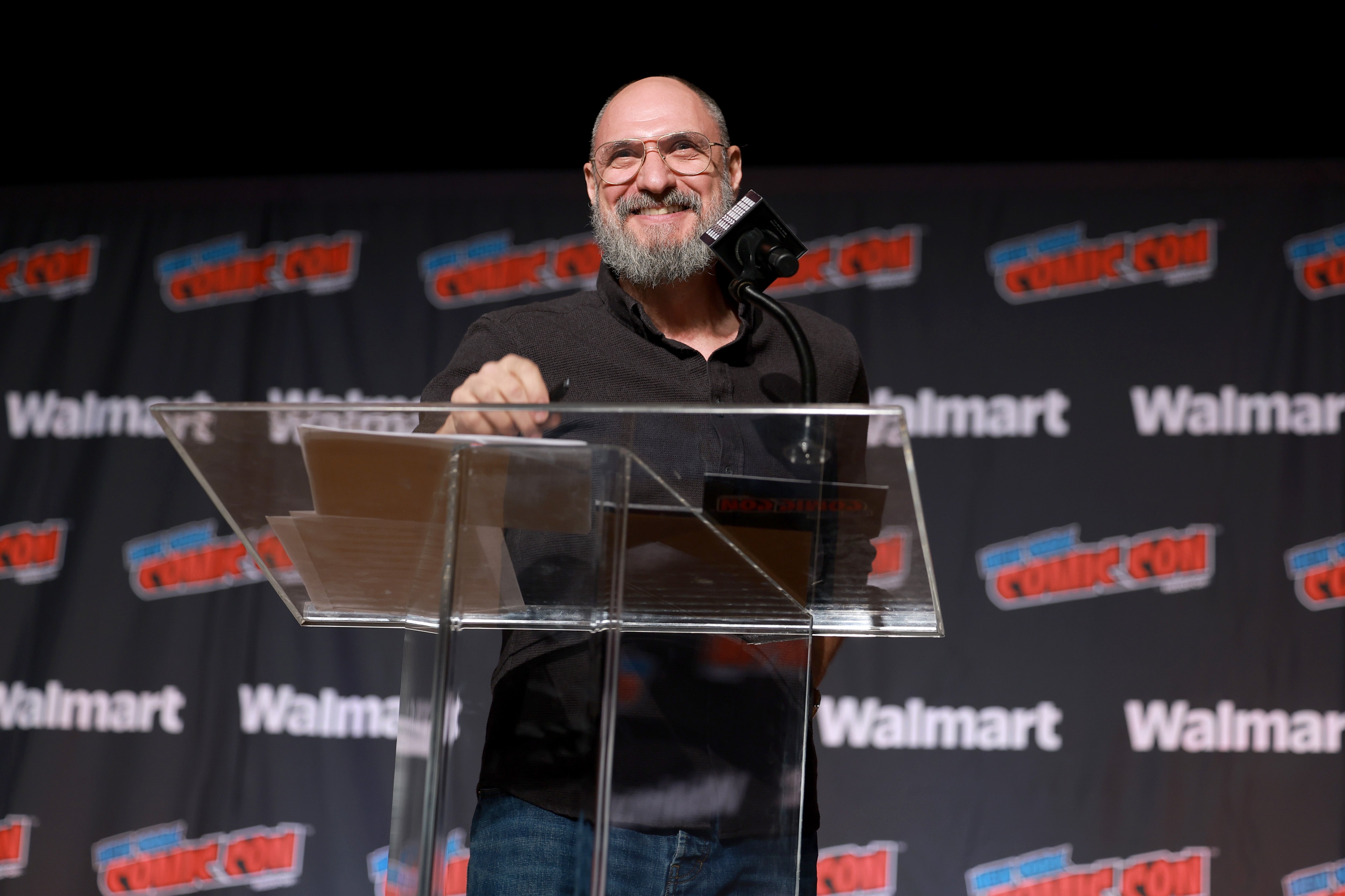Man speaking at a podium at a comic convention, with a backdrop showing logos for Walmart and Comic Con