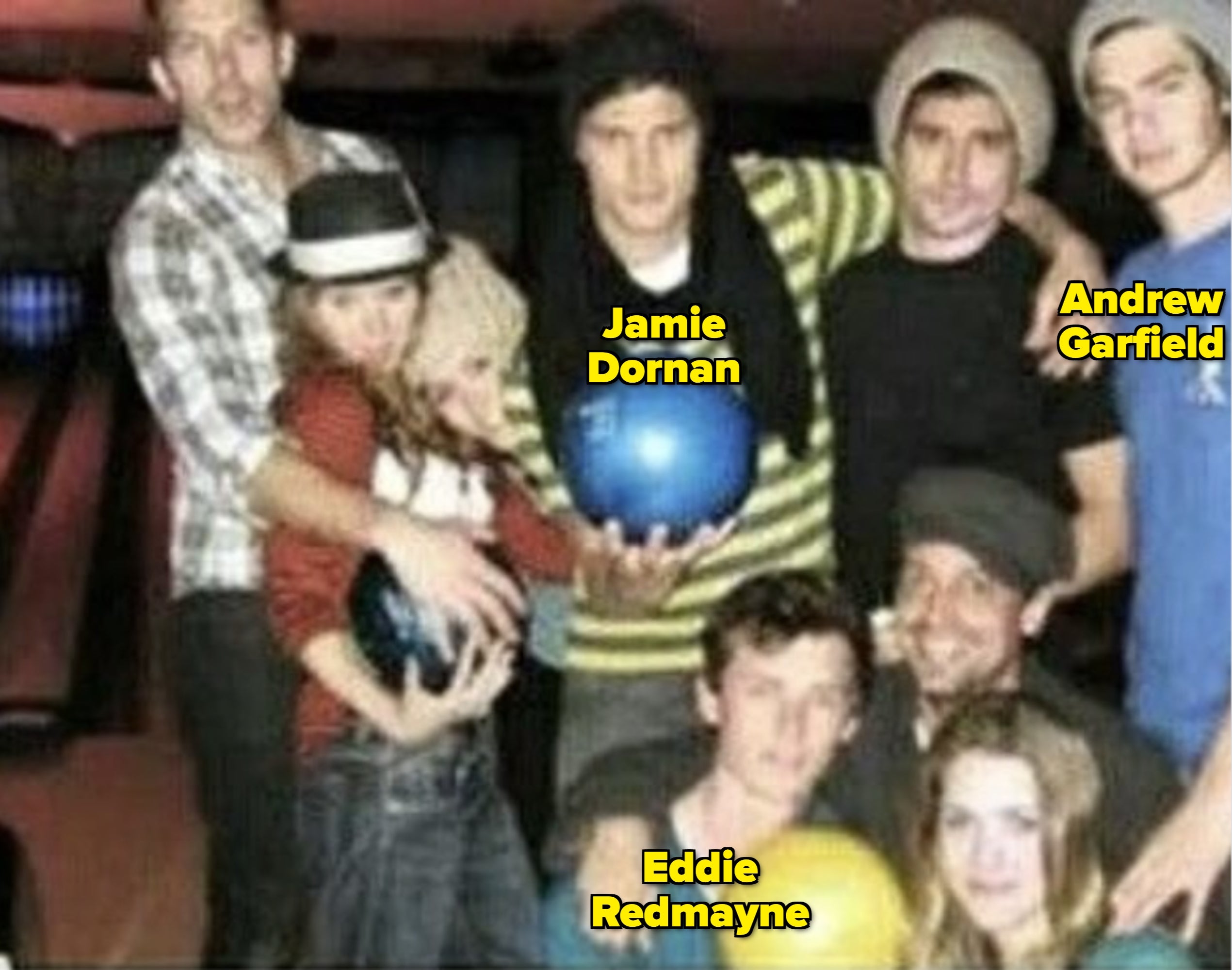 Group of people at a bowling alley, holding bowling balls, dressed casually in hats and scarves, smiling and posing for a photo
