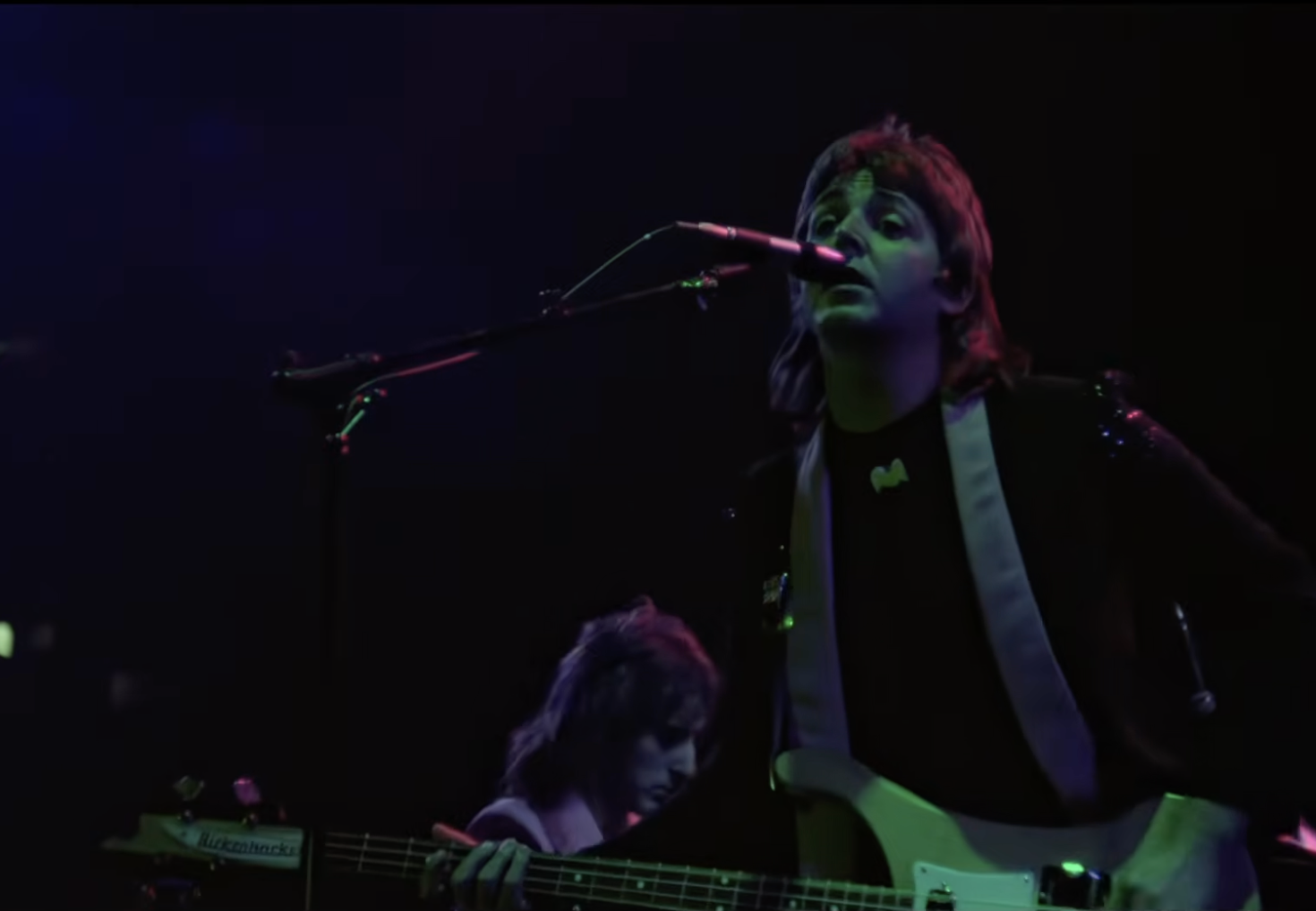Musician playing a guitar and singing on stage with a band in the background during a live concert performance