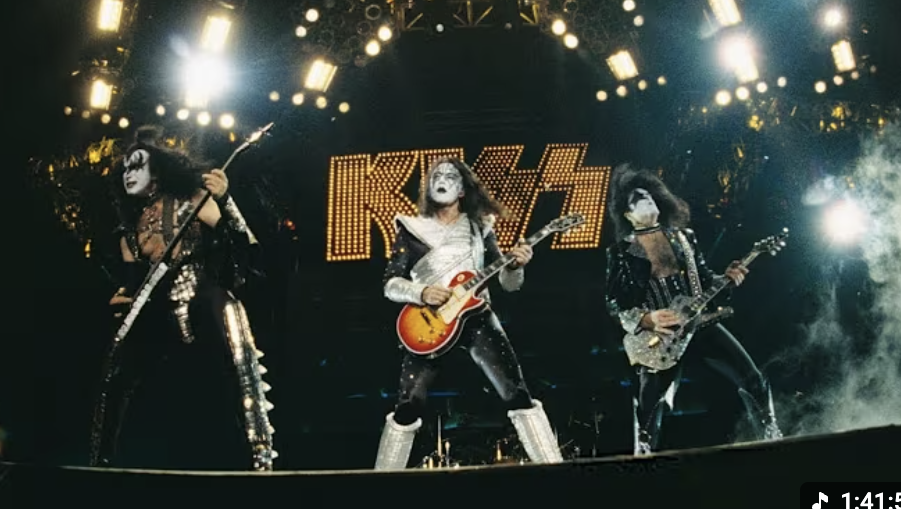 Rock band KISS performing on stage with signature makeup and costumes, energetic poses, and bright stage lights