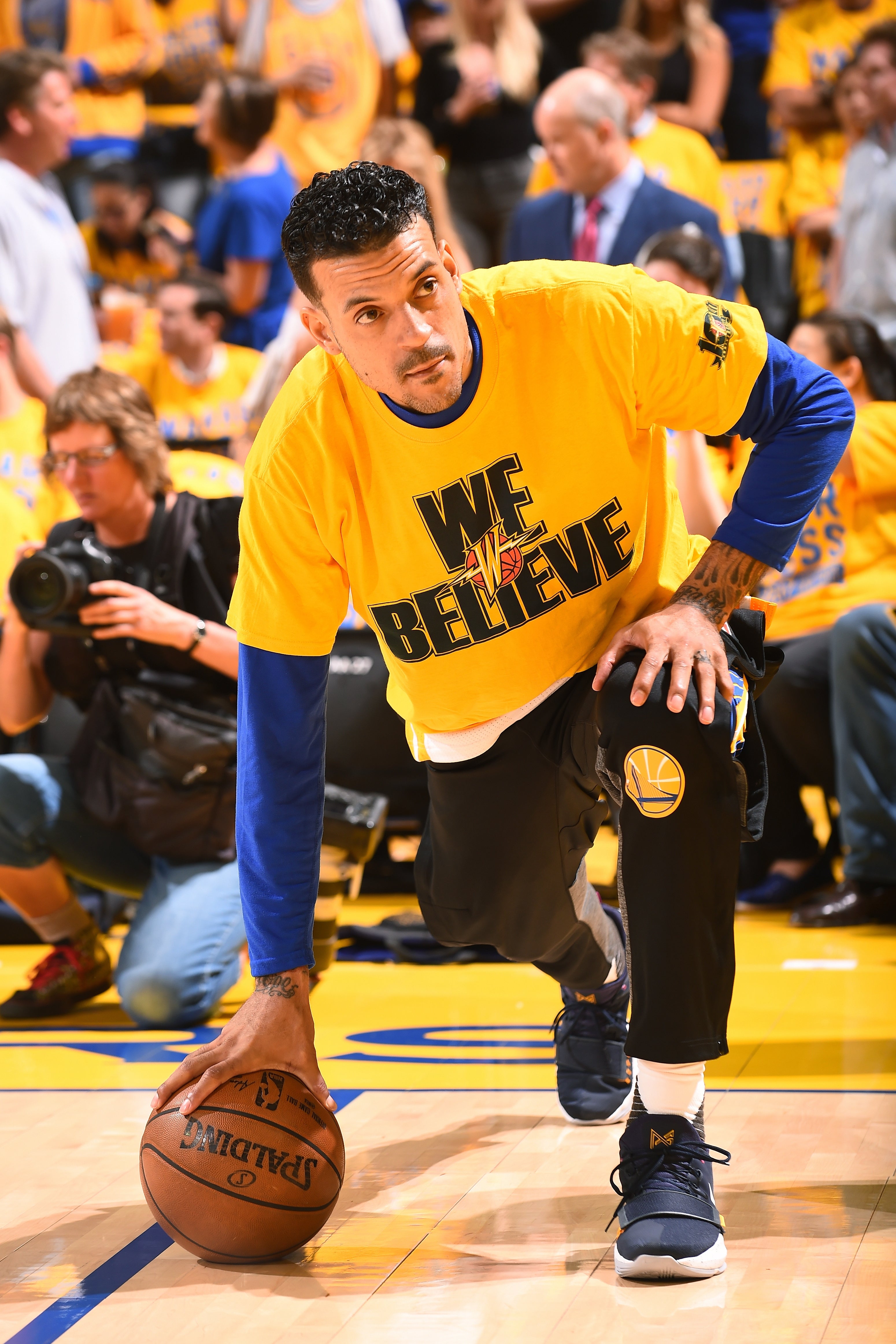 Person kneeling on a basketball court wearing a "We Believe" shirt, preparing to play basketball