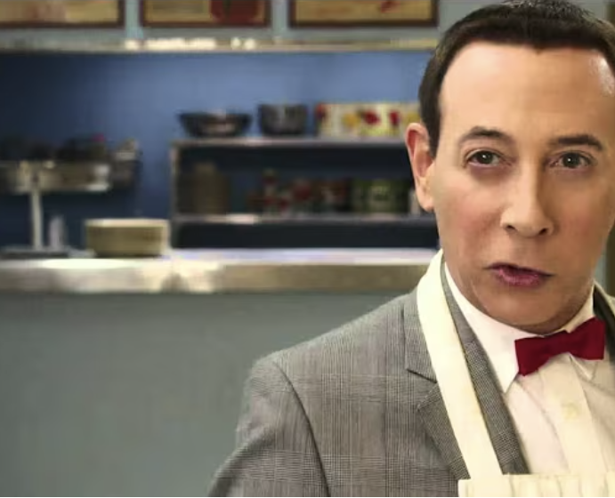 Person in a suit with a red bow tie stands in a diner setting, wearing an apron