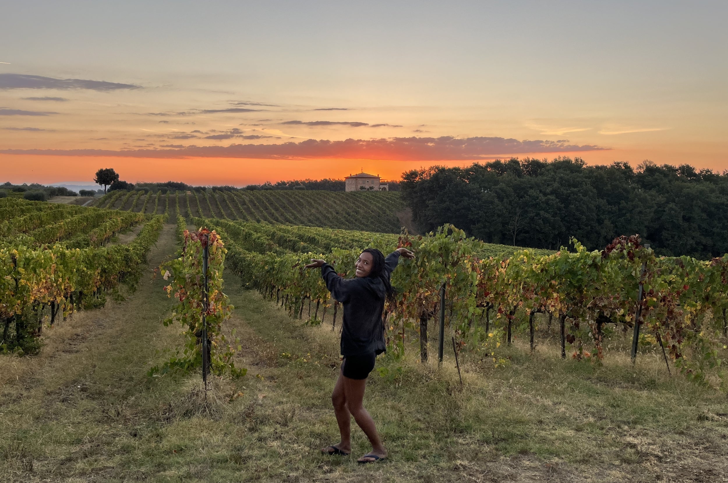 Person posing playfully in a vineyard at sunset, with rows of grapevines and a distant house in the background