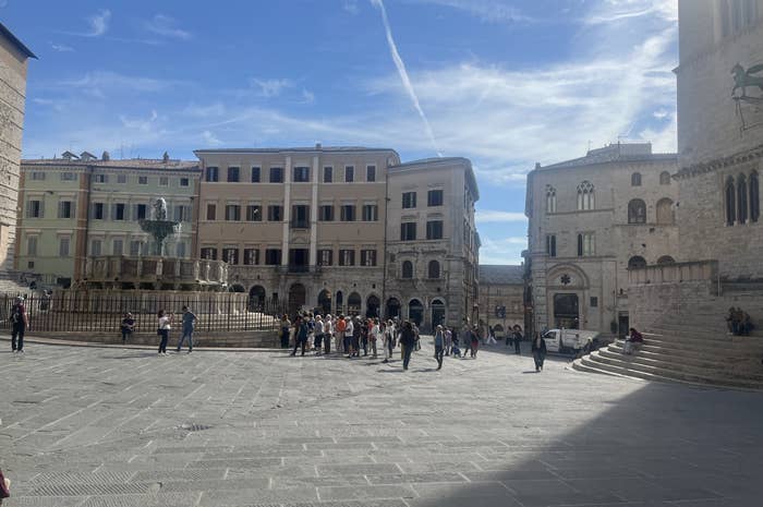People gather in a historic European plaza with a central fountain and surrounding old buildings