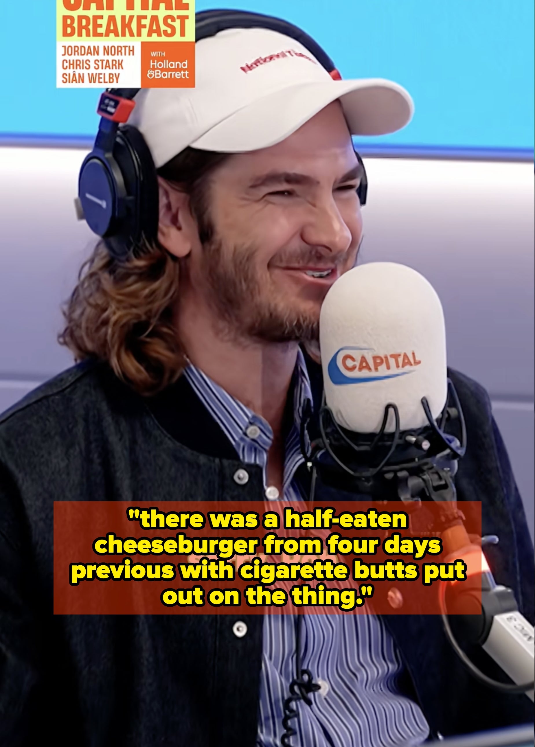 Person with long hair and beard wearing a striped shirt, jacket, and a cap, speaking into a microphone labeled "Capital." Studio background