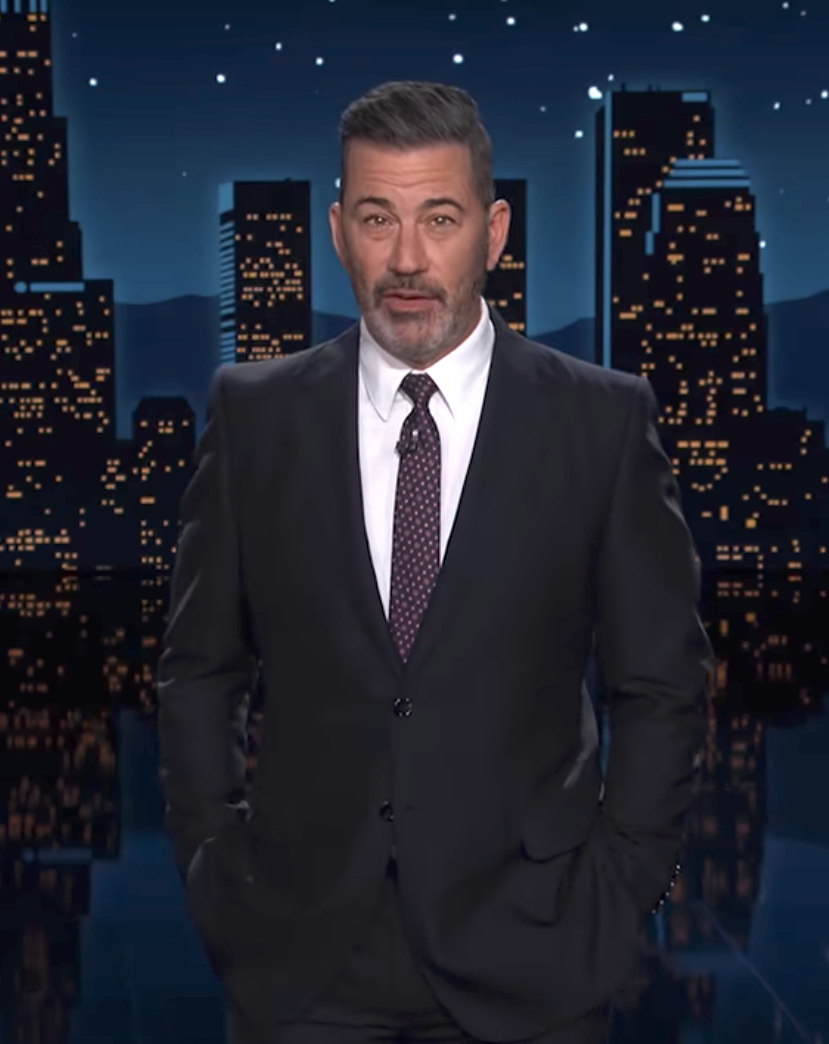 A person in a suit stands against a nighttime cityscape backdrop with stars, possibly during a television show segment
