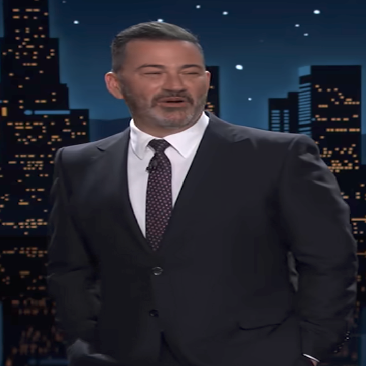 Person in a suit on a cityscape-themed talk show set, standing with hands in pockets