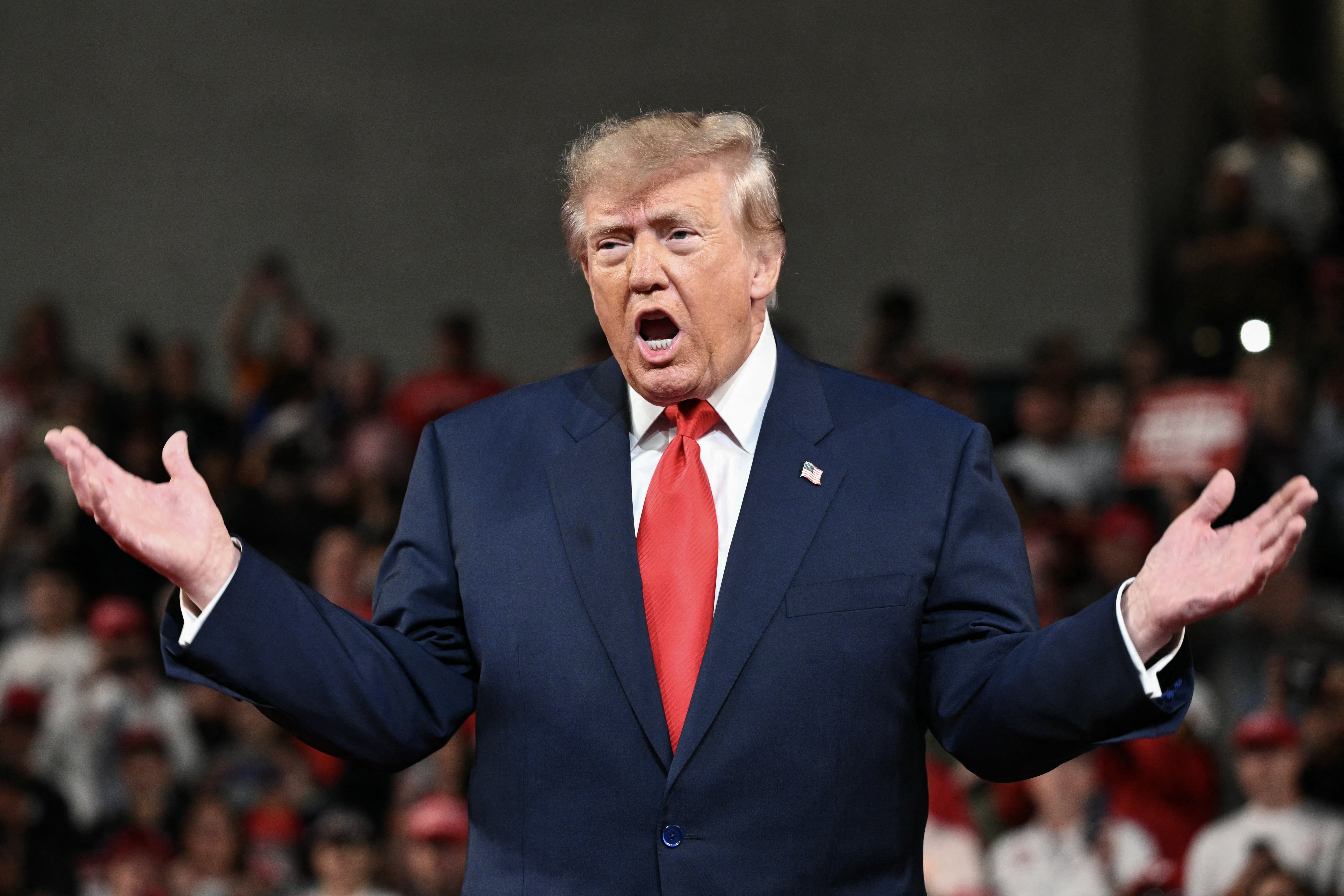 A person in a suit with a red tie gestures with open hands, speaking energetically at a public event with a crowd in the background