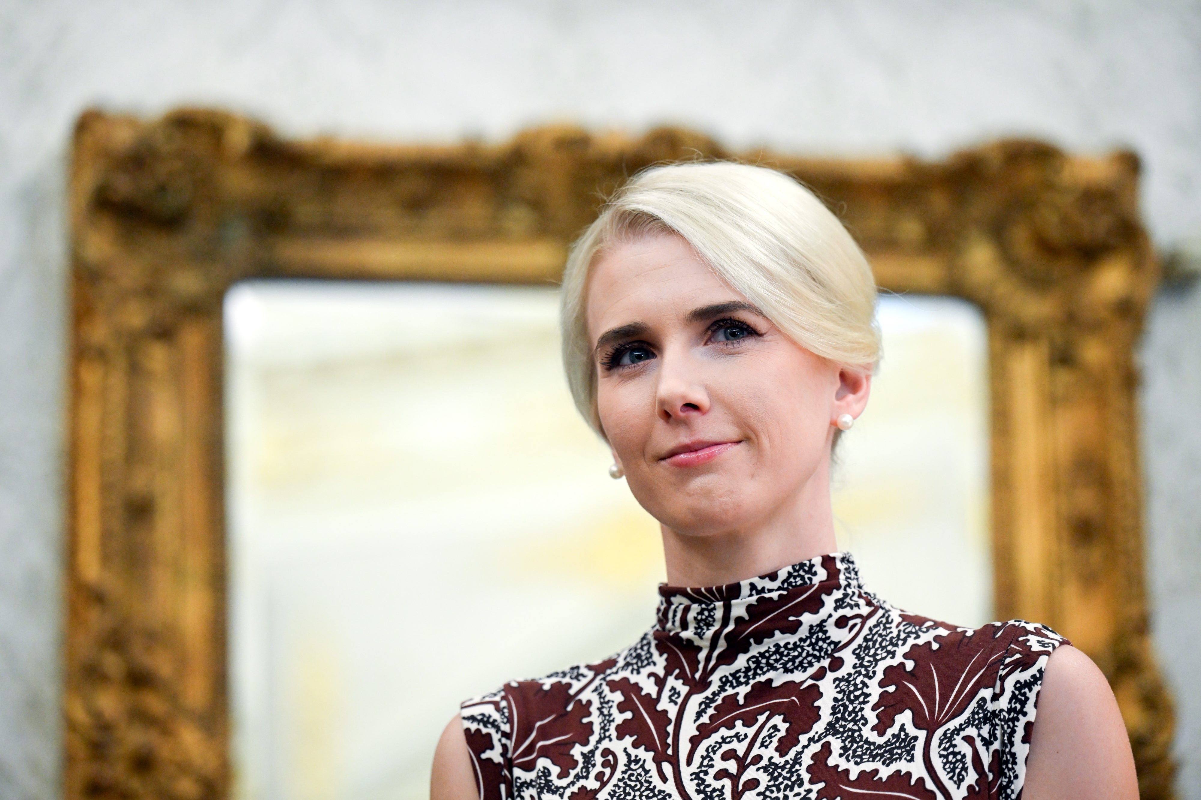 A woman with short blonde hair in a patterned, high-neck dress stands in front of an ornate mirror