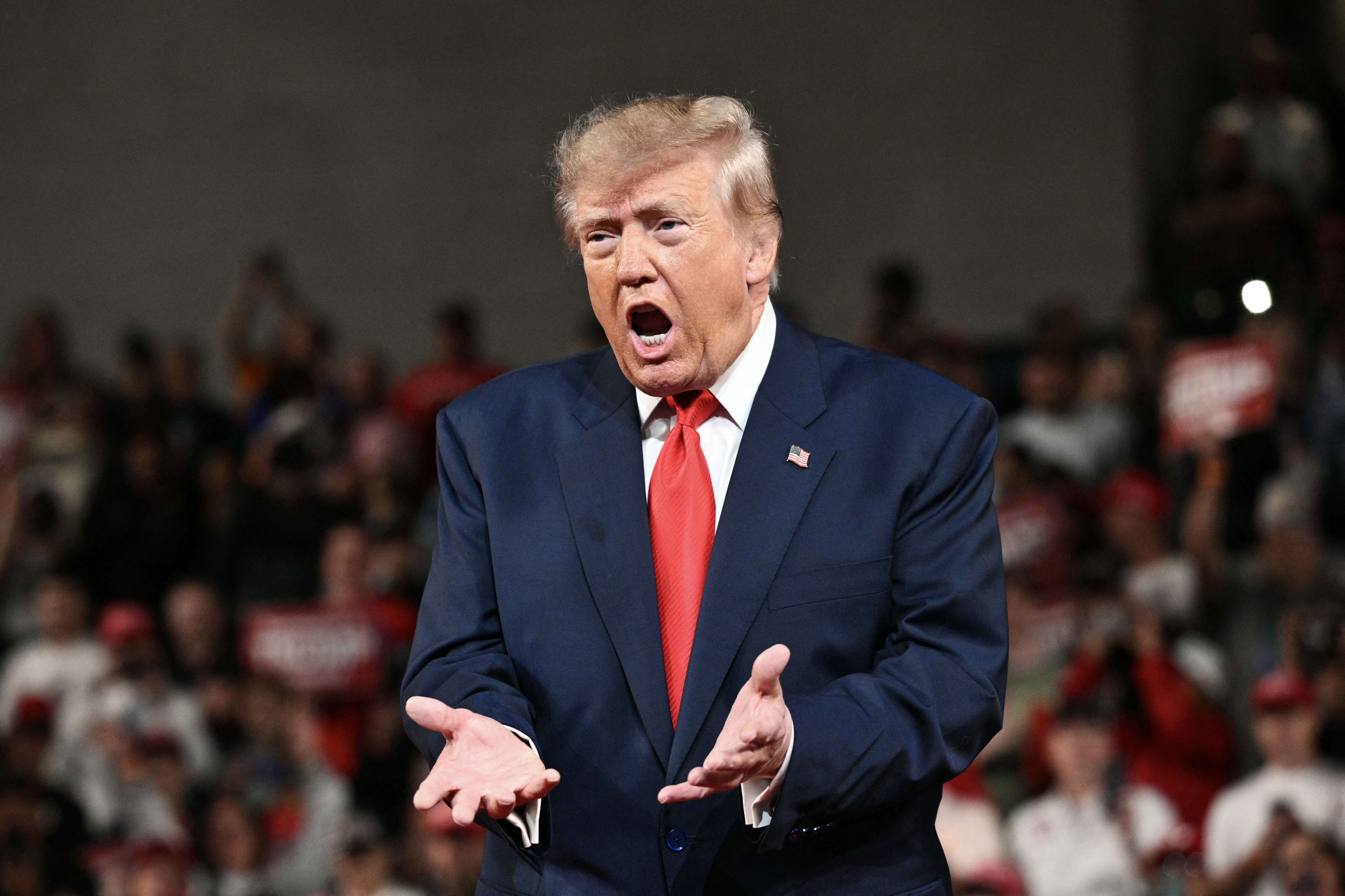 A person in a suit and tie gestures emphatically, speaking to a crowd at an event