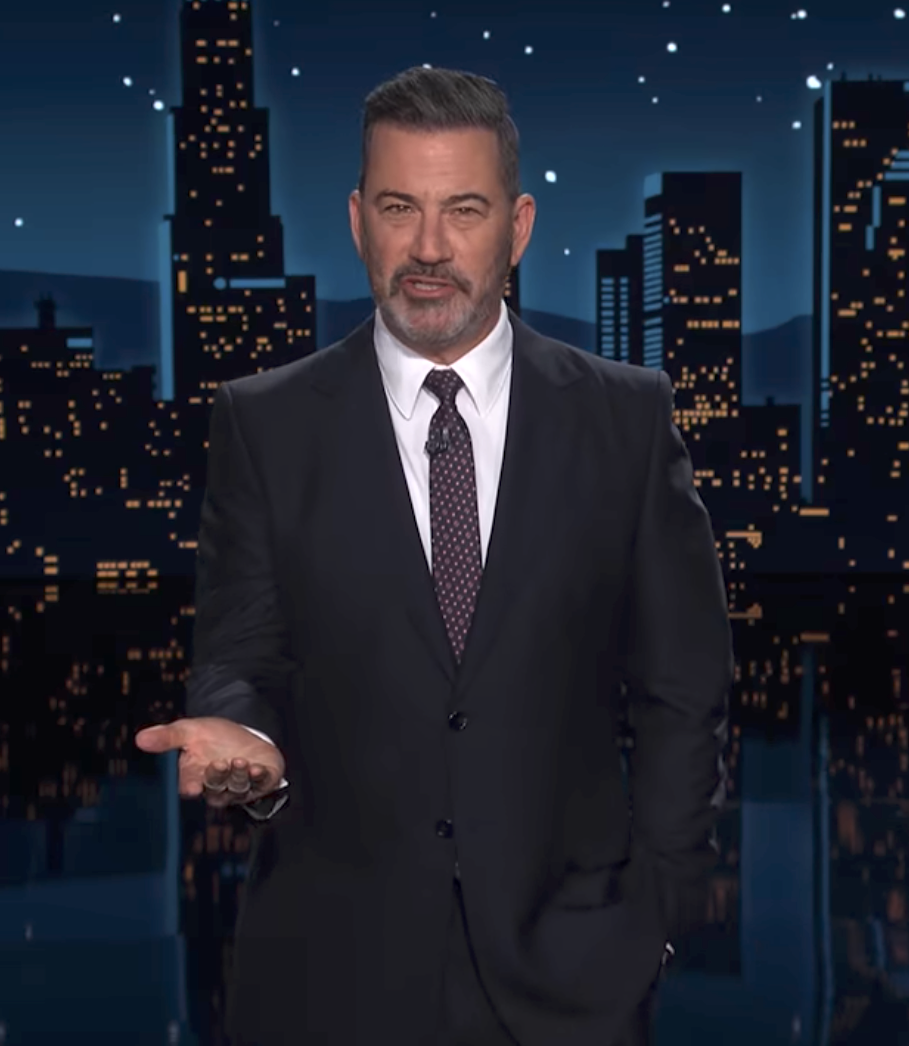 Man in a suit stands on a stage with a cityscape backdrop, gesturing with one hand