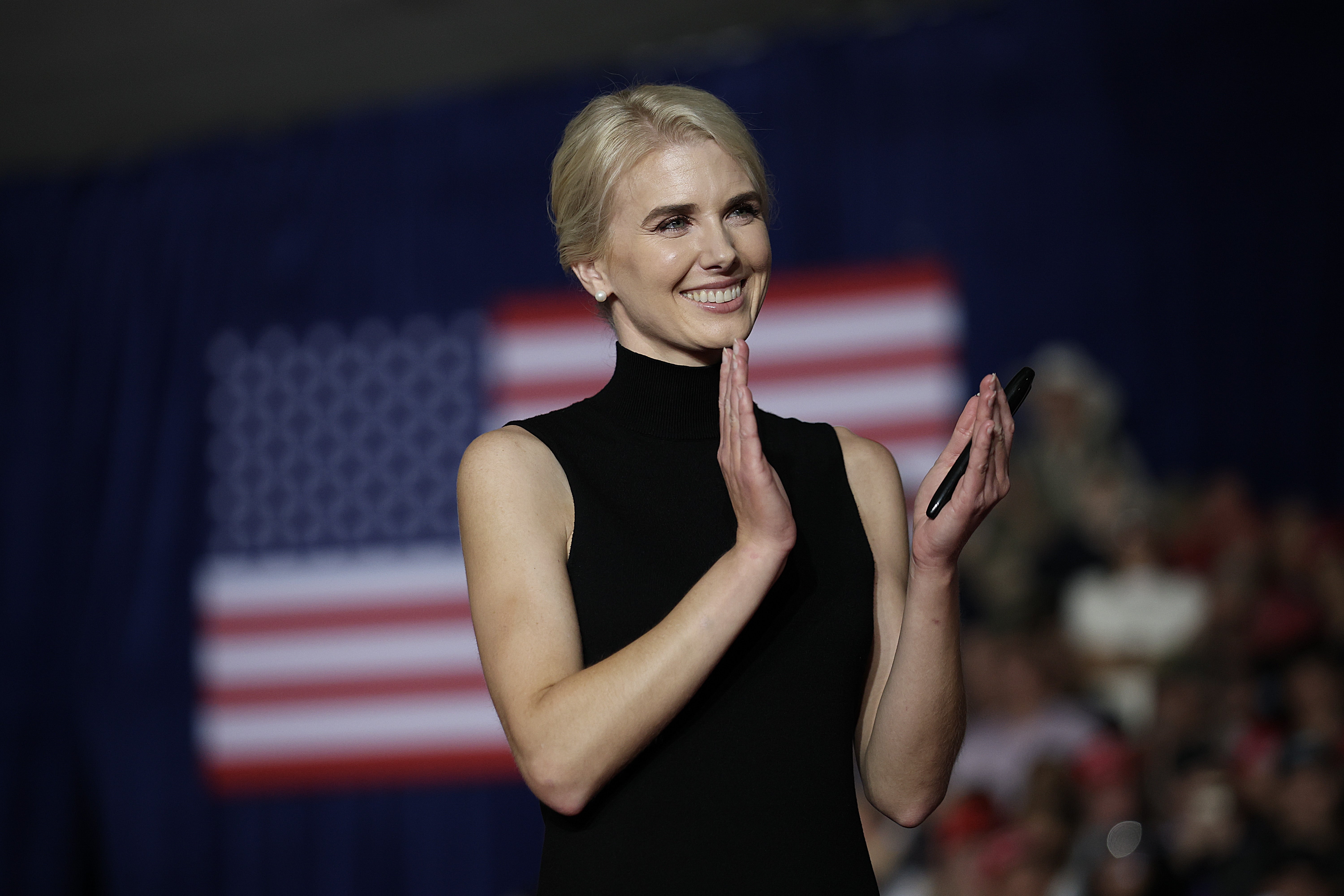 Person in a sleeveless black dress clapping on stage, with a U.S. flag blurred in the background